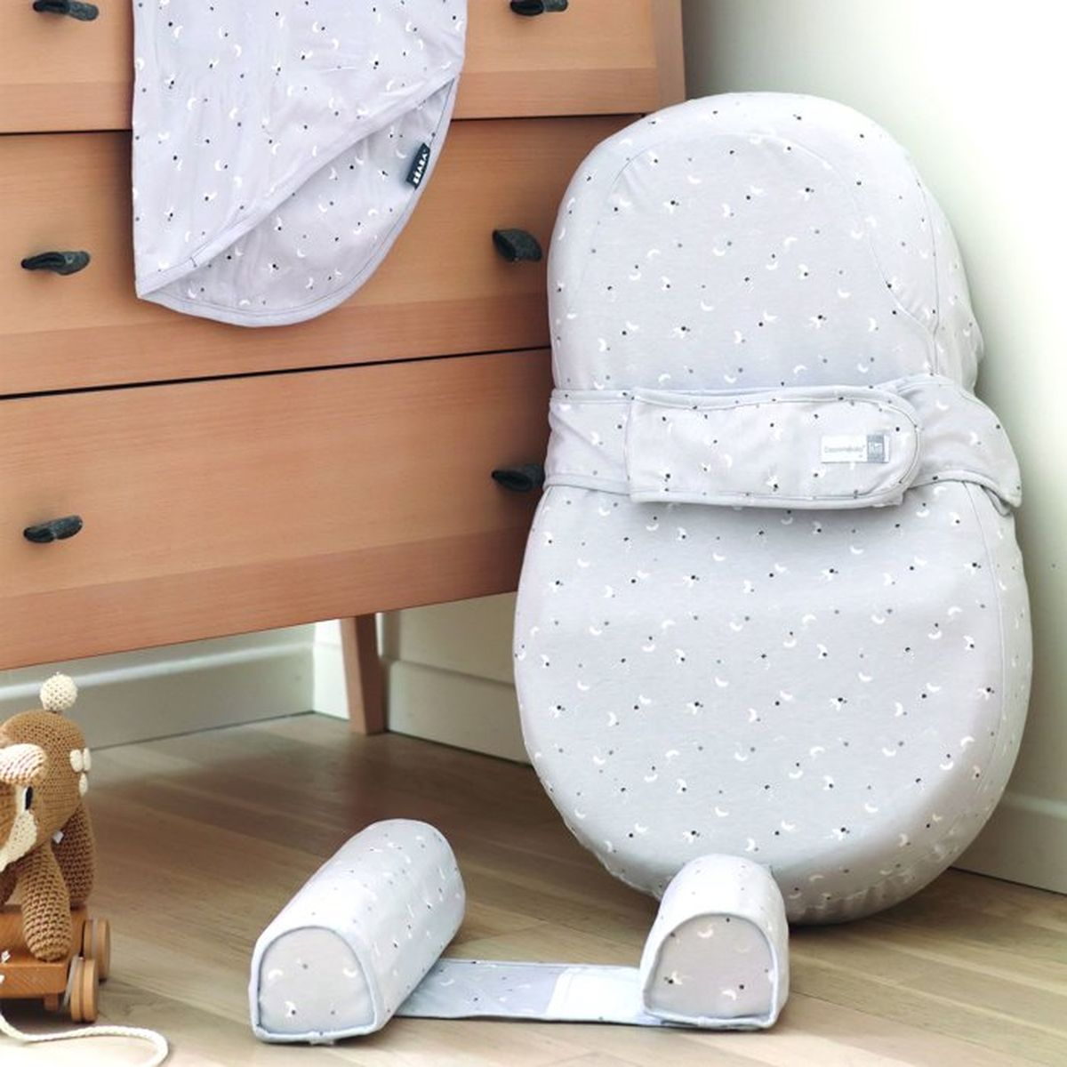 Ein graues Babynestchen mit Mondmuster steht auf einem Holzboden vor einer Kommode, daneben ein Stricktier.
