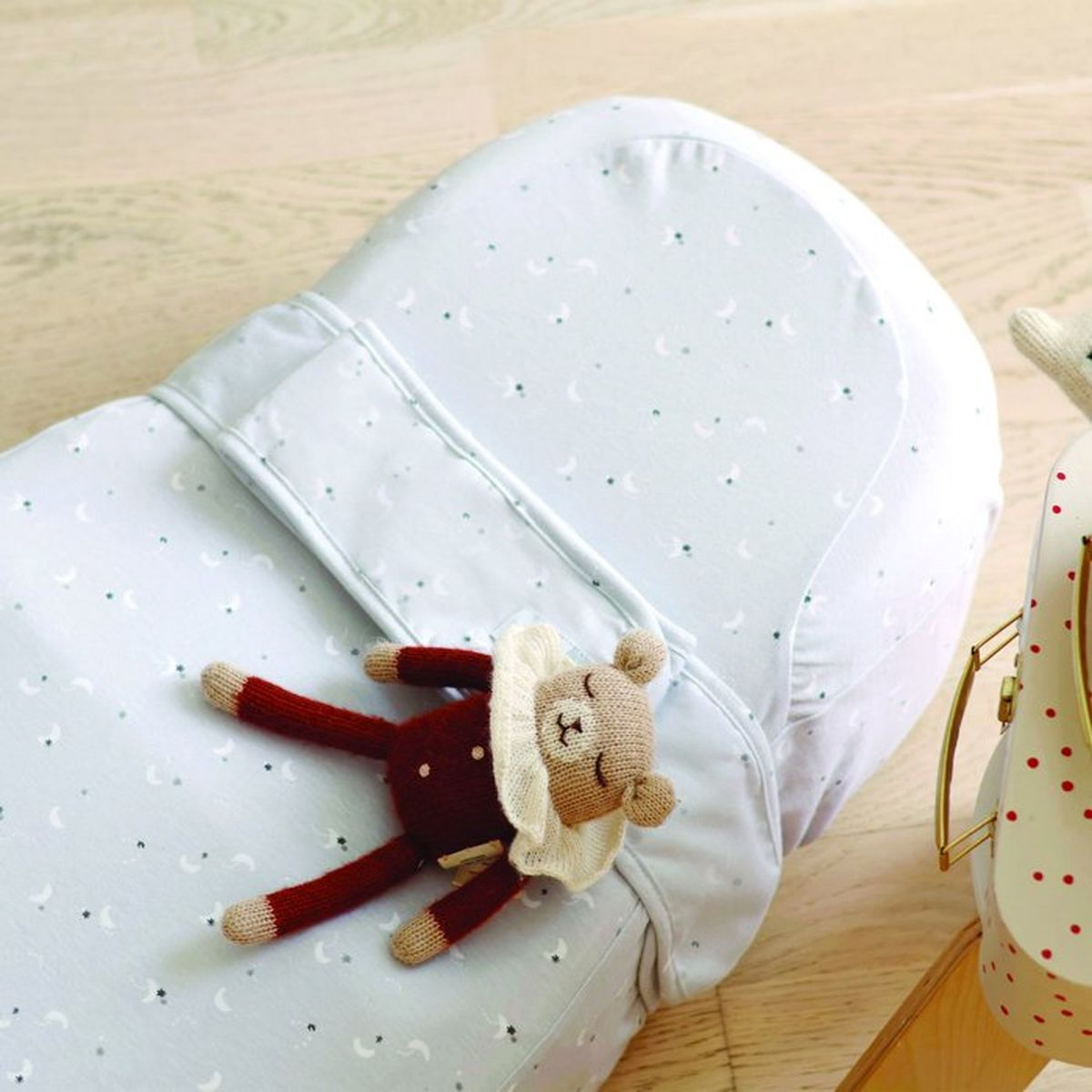 Ein Kuscheltier-Bärchen liegt auf einer hellblauen Babyliege mit Mond- und Sternenmuster auf Holzboden.