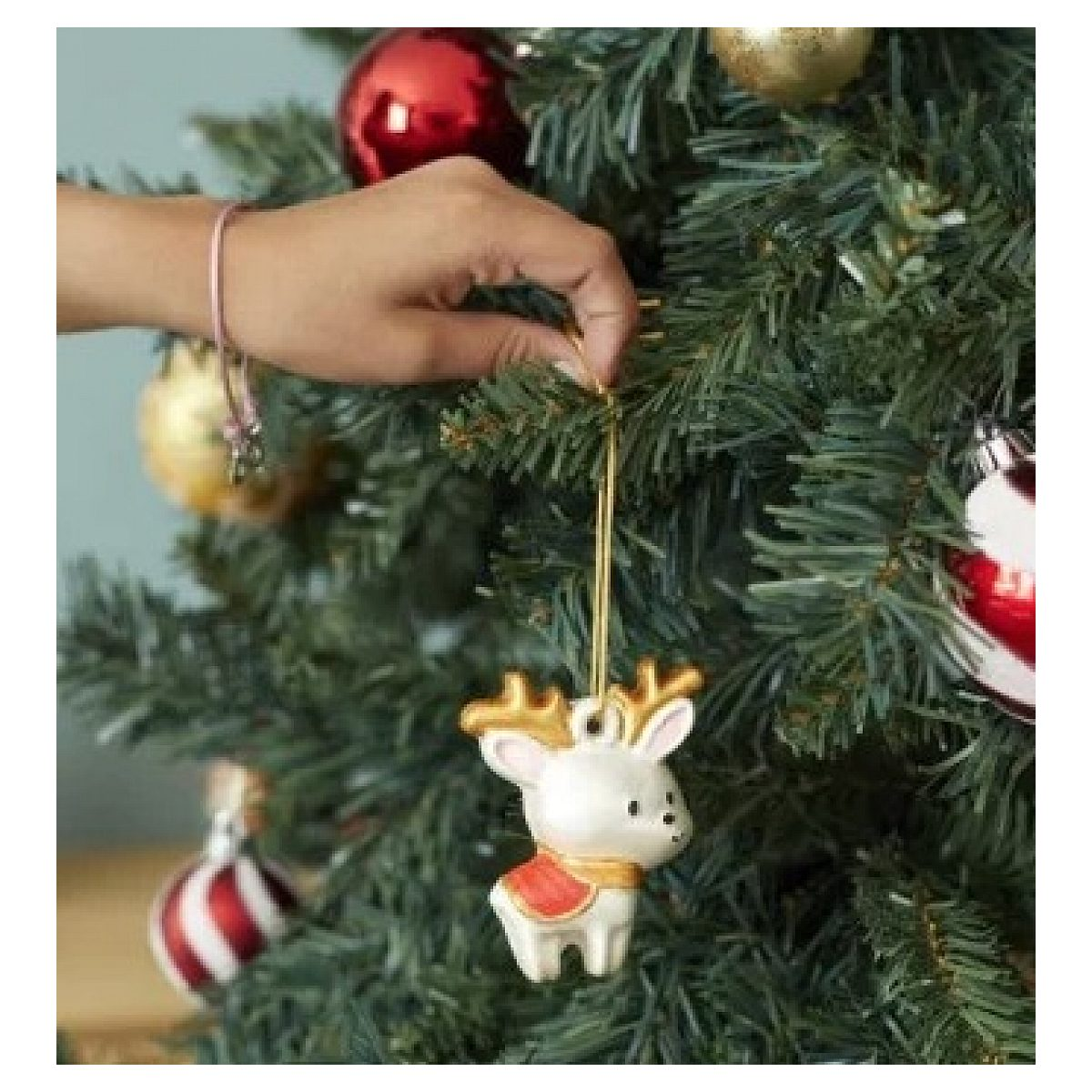 Eine Hand hängt ein kleines weiß-goldenes Rentier-Ornament an einen Weihnachtsbaum.