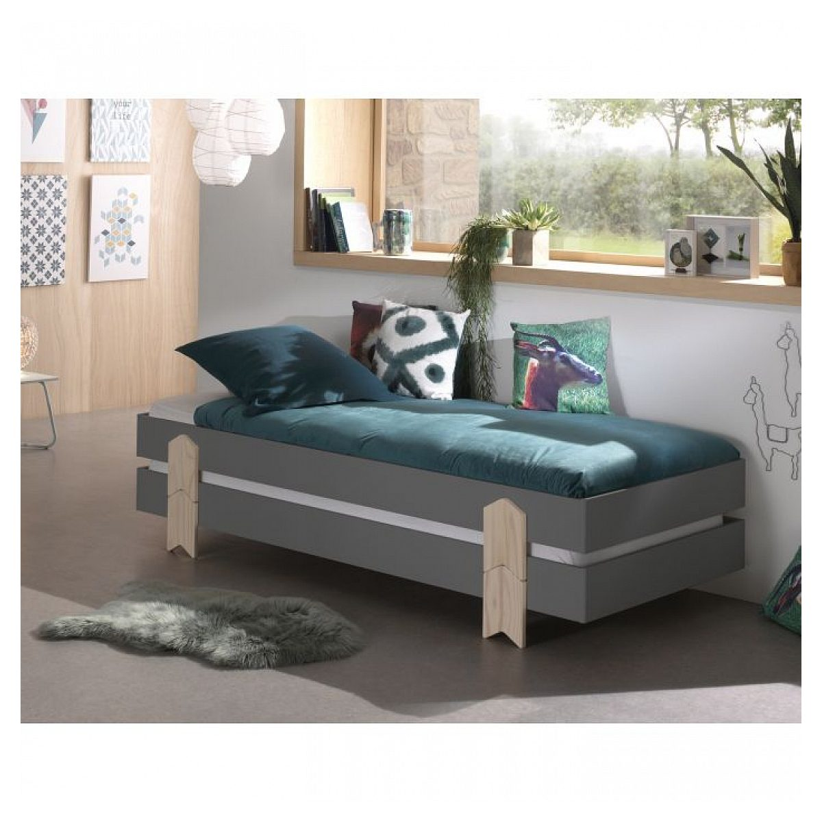 Ein graues Bett mit hellen Holzbeinen steht in einem hellen Zimmer vor einem Fenster mit Pflanzen.