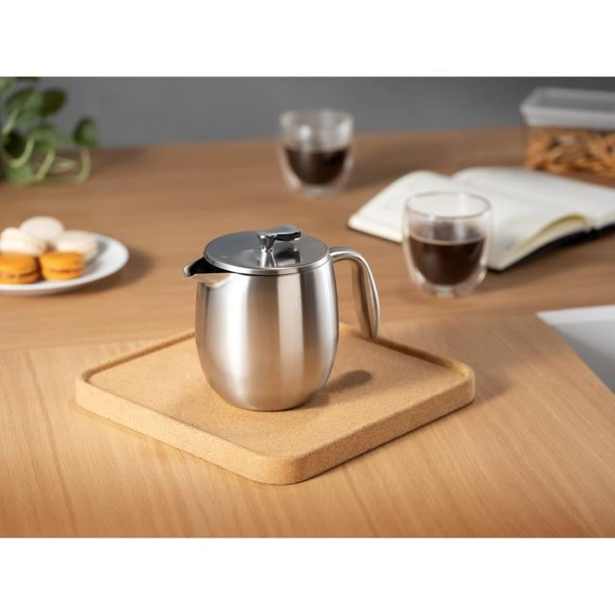 Eine silberne Kaffeekanne auf einem Korktablett auf einem Holztisch mit Gläsern, Buch und Gebäck im Hintergrund.