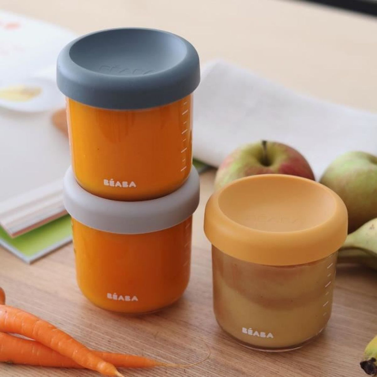Zwei stapelbare und ein einzelner Behälter mit orangem Inhalt und bunten Deckeln auf einem Holztisch, umgeben von Obst.