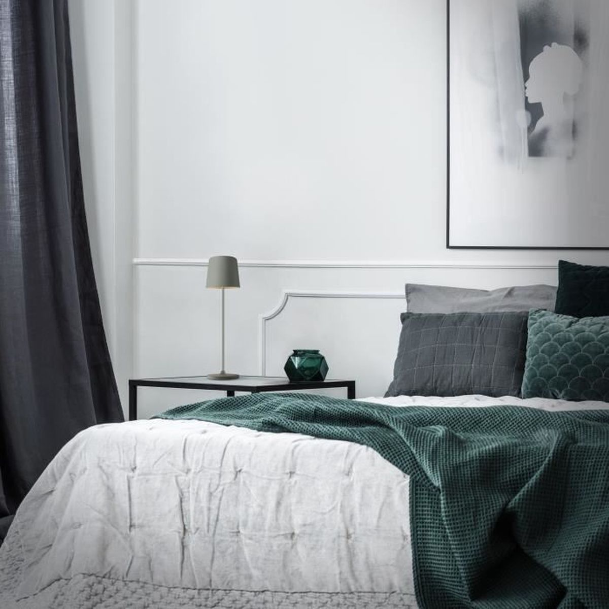 Ein minimalistisches Schlafzimmer mit weißer Wand, dunklem Vorhang, einem Bett mit grüner Decke und Kissen sowie einer Lampe.