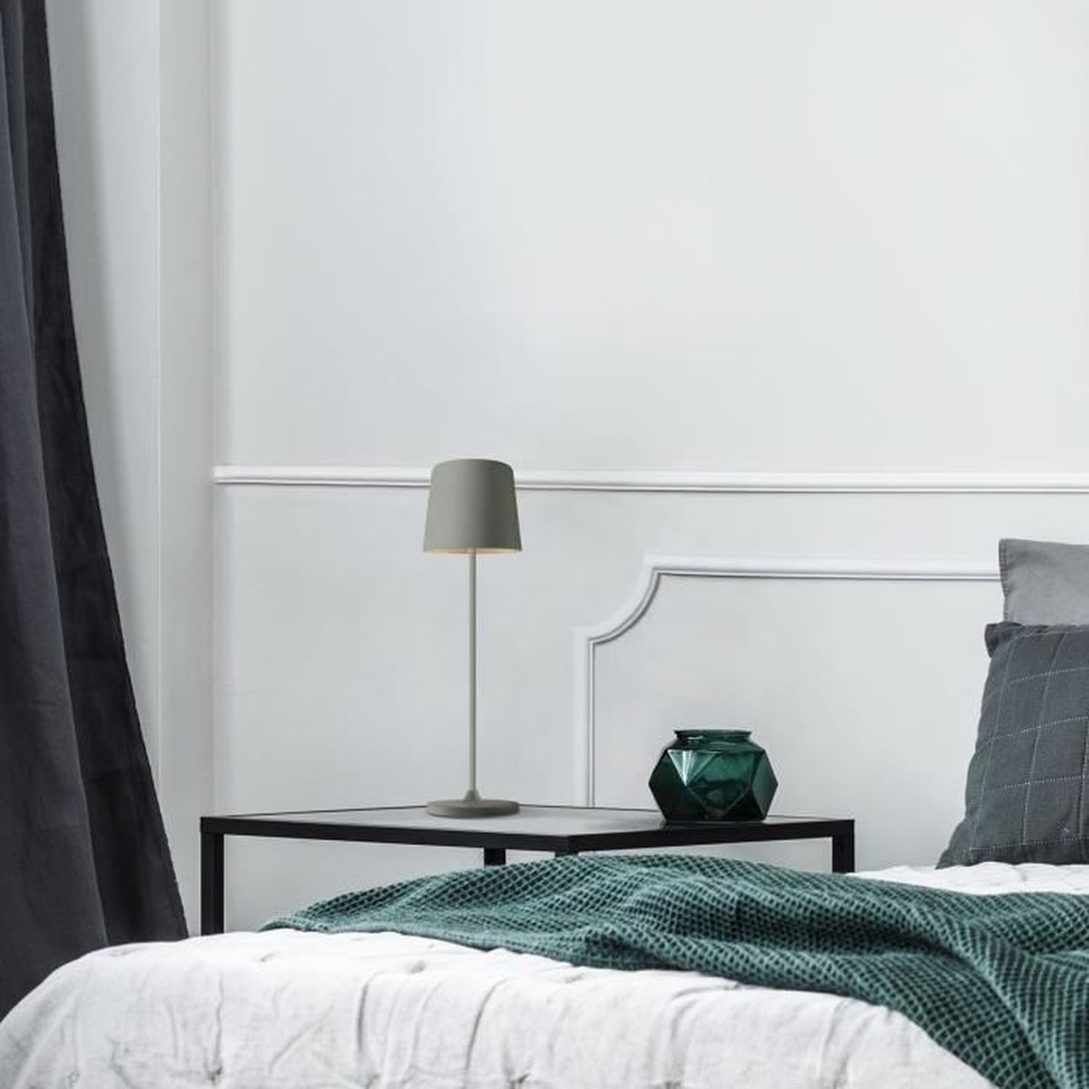 Eine graue Lampe und eine grüne Vase stehen auf einem Nachttisch neben einem Bett.