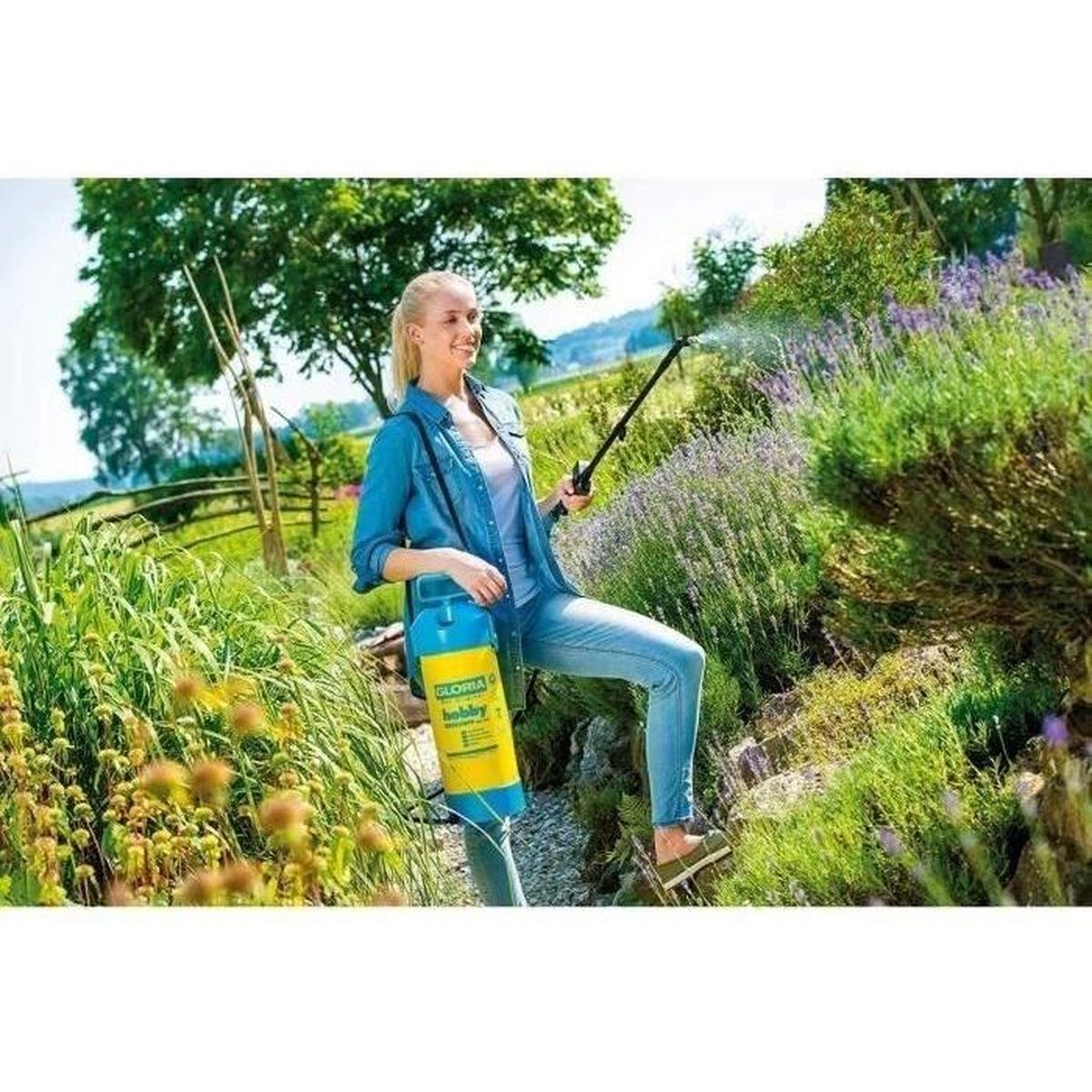 Eine Frau in Jeans sprüht Lavendelpflanzen in einem sonnigen Garten mit einem gelb-blauen Sprühgerät.