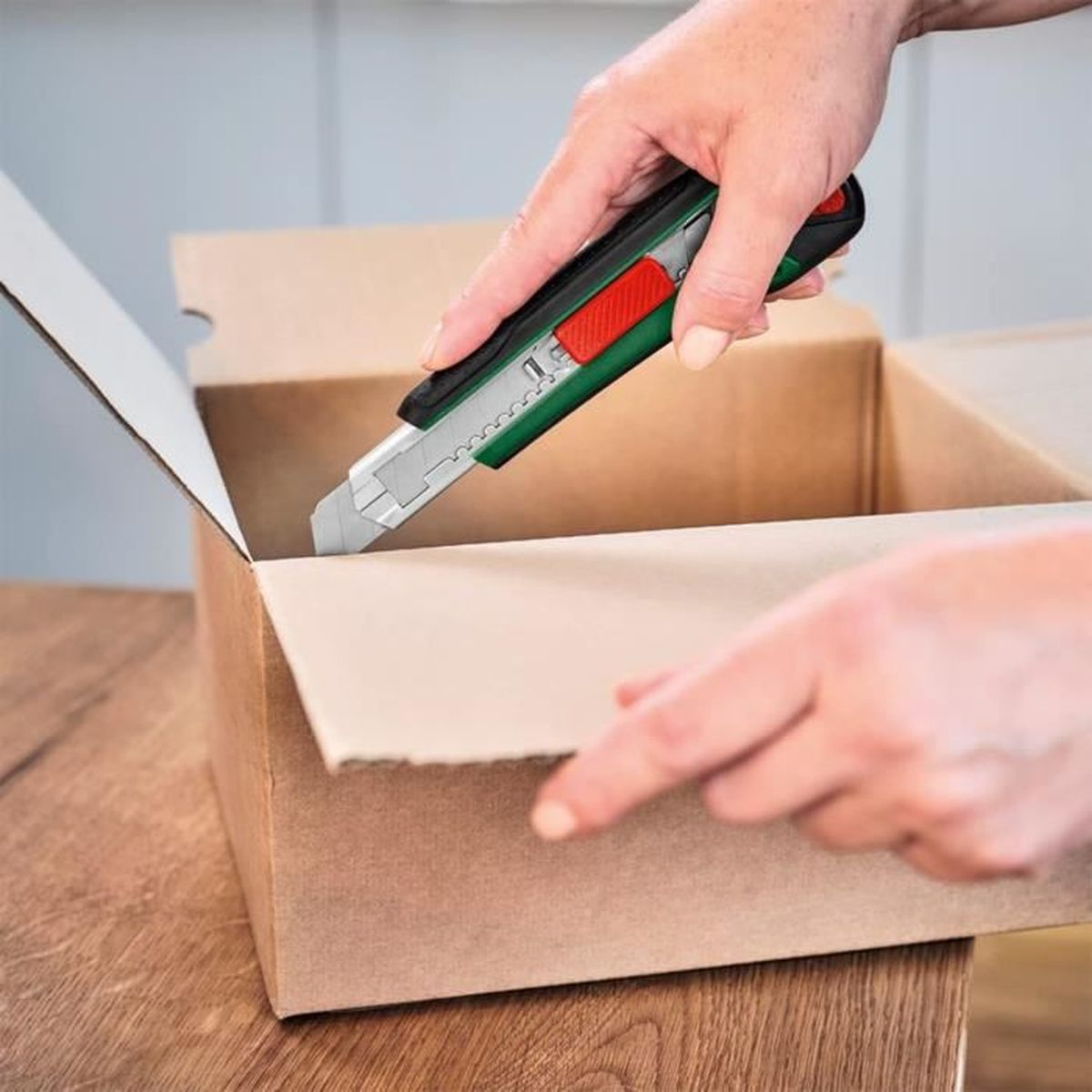 Ein Paar Hände öffnet mit einem grünen Teppichmesser vorsichtig einen braunen Karton auf einem Holztisch.
