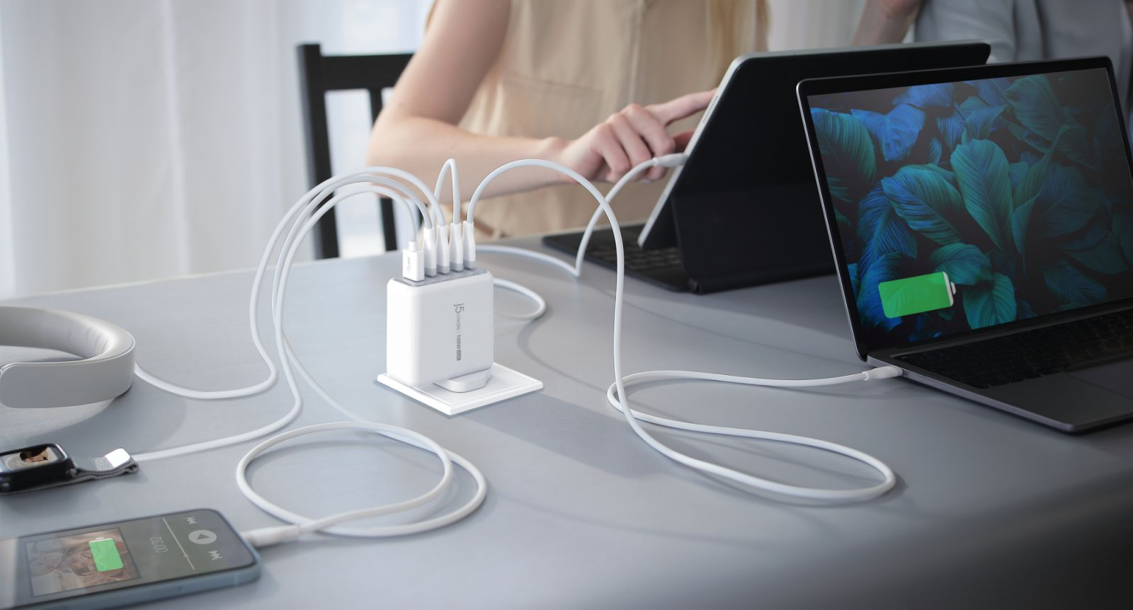 Ein weißes USB-C-Ladegerät auf einem Tisch verbindet weiße Kabel mit einem Laptop, Smartphone und Tablet.