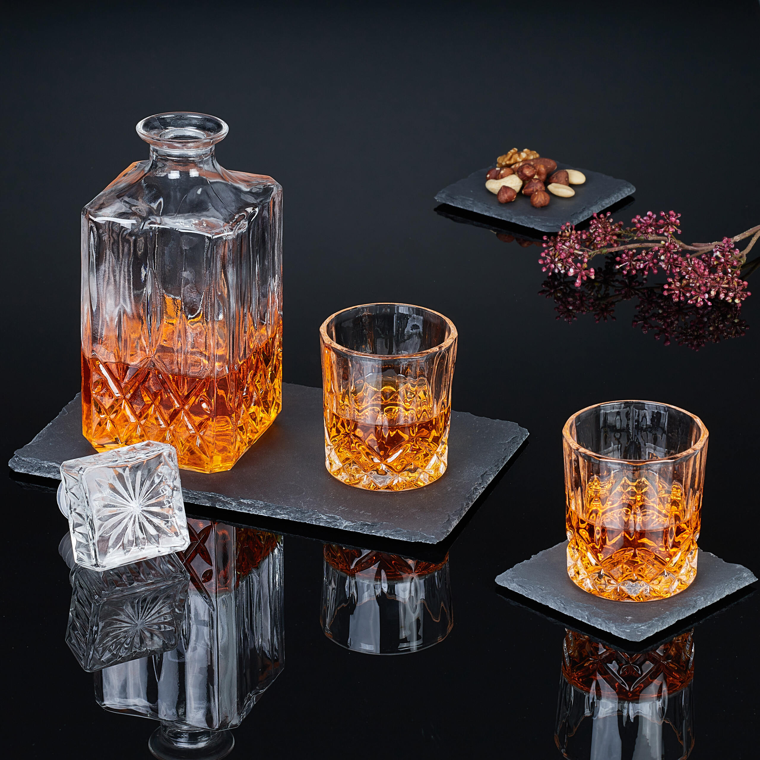 Whiskeykaraffe und zwei Gläser mit Bernstein-Whiskey auf dunklen Schieferplatten, begleitet von Nüssen und Blüten.