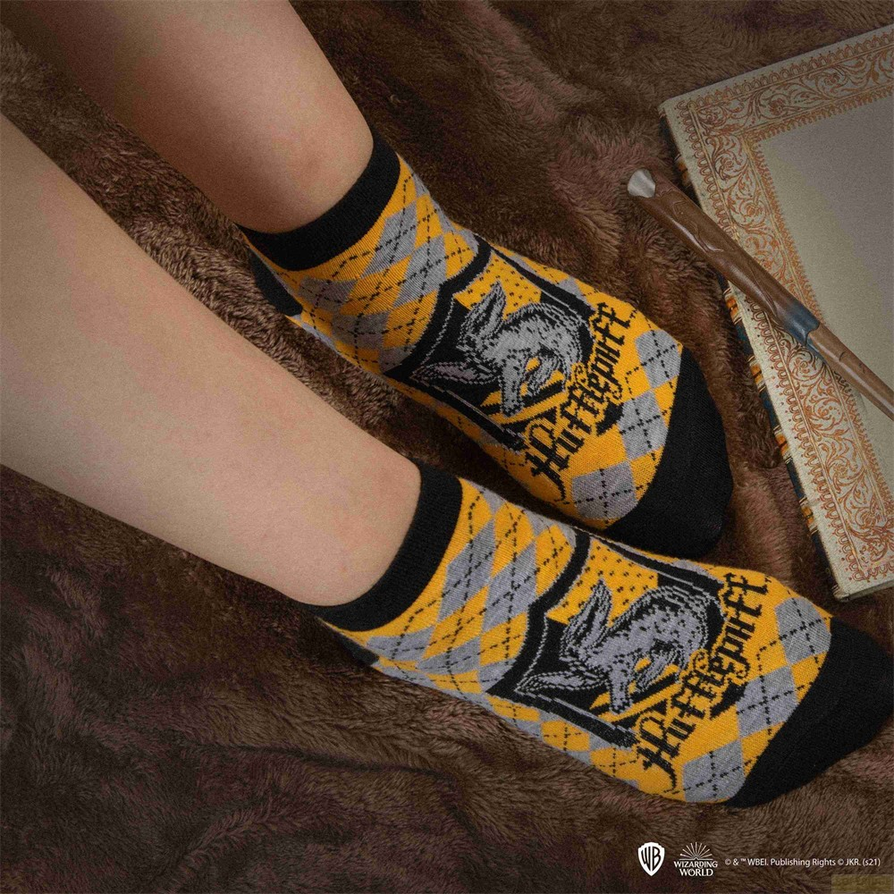 Persona lleva calcetines Hufflepuff de rombos, con varita y libro sobre manta marrón cerca.