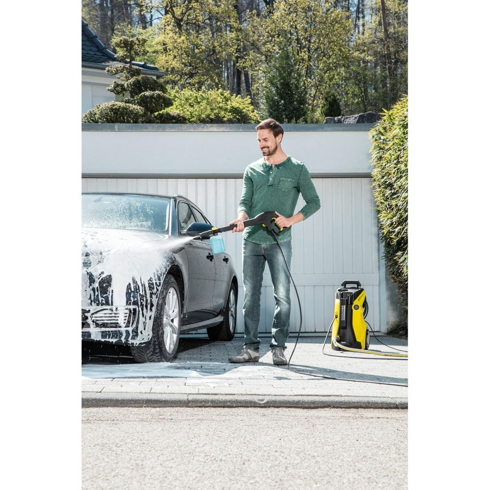 Ein Mann wäscht ein schwarzes Auto mit einem gelben Hochdruckreiniger vor einer Garage.