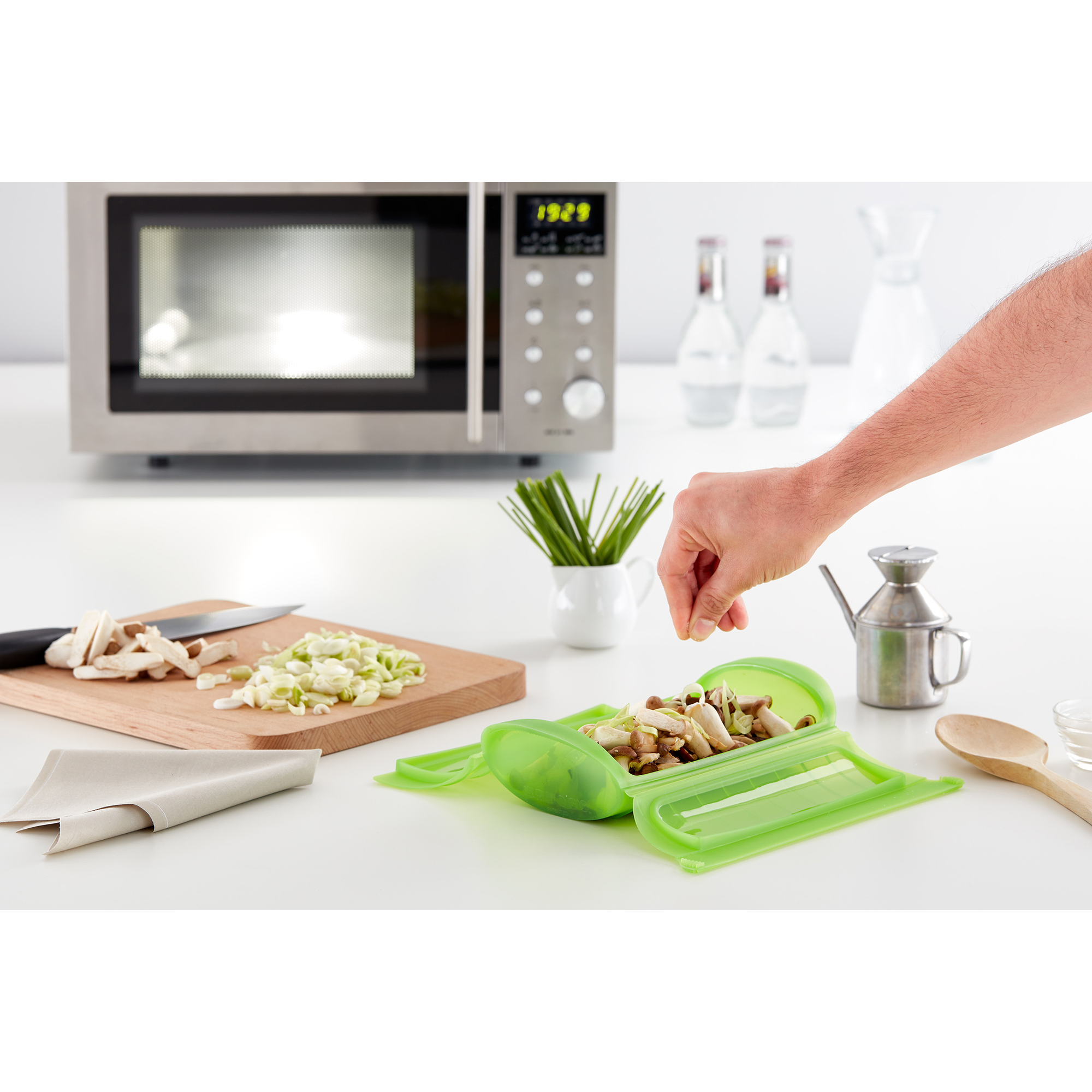 Eine Hand gibt Zutaten in eine grüne Silikon-Dampfgarform auf einer weißen Küchentheke vor einer Mikrowelle.