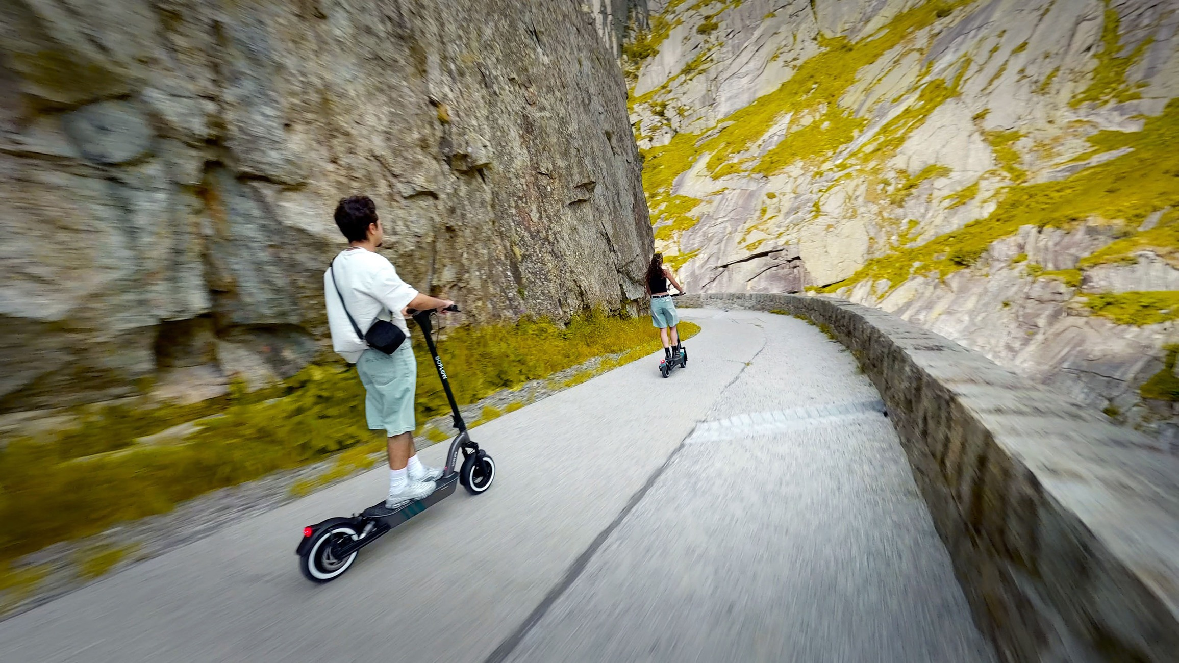 Zwei Personen fahren auf E-Scootern eine kurvige, von hohen Felsen gesäumte Straße entlang.