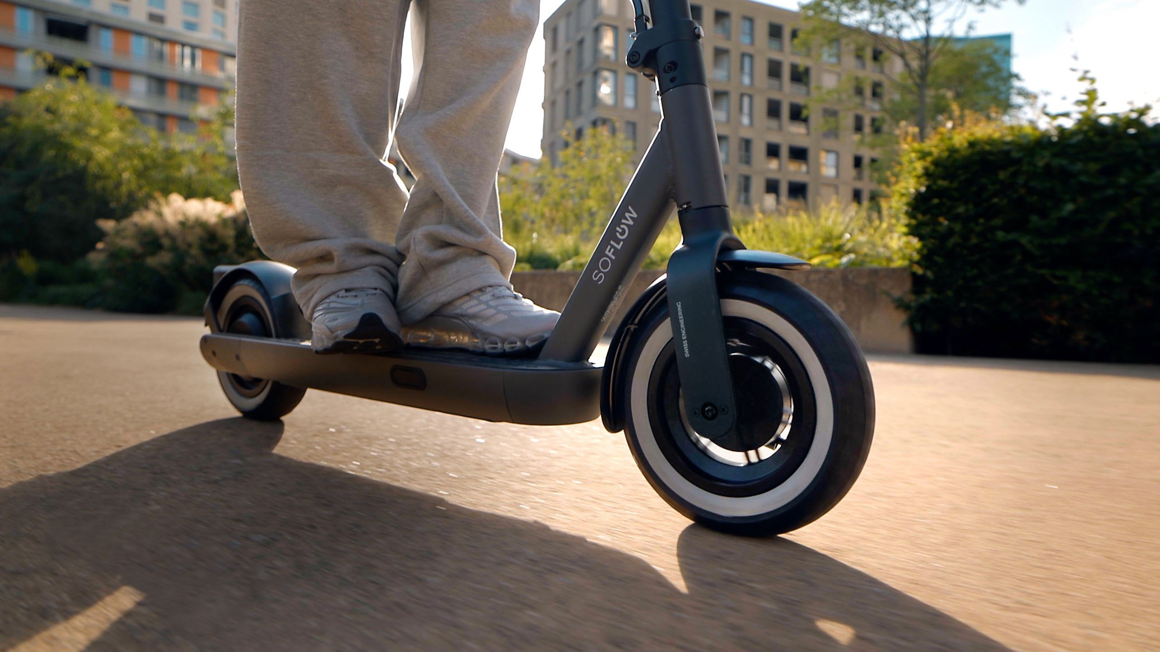Eine Person steht auf einem grauen SoFlow E-Scooter, der im Sonnenschein auf einem Weg fährt.
