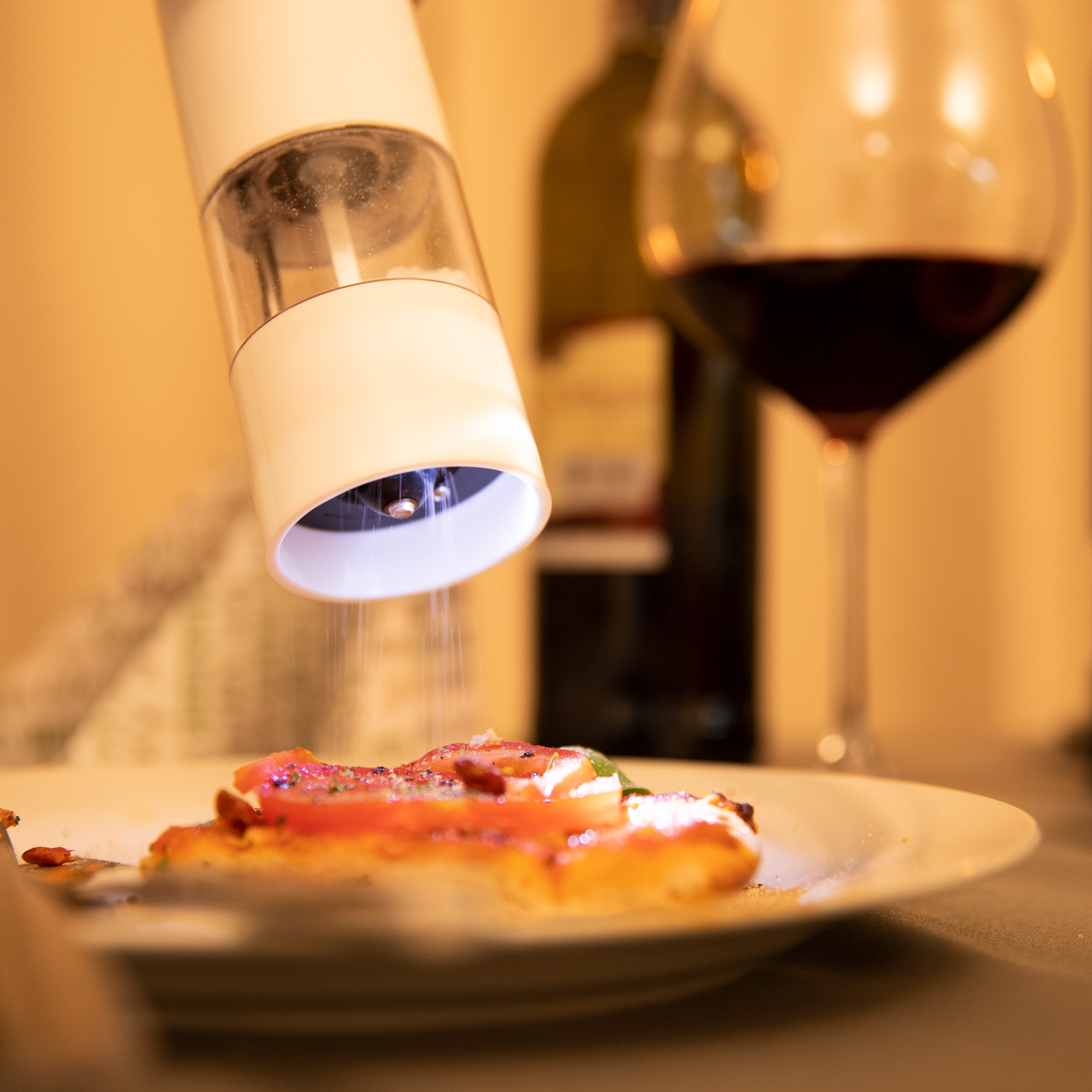 Eine elektrische Mühle streut Gewürze auf eine Pizza mit Tomaten; im Hintergrund stehen Wein und ein Glas.