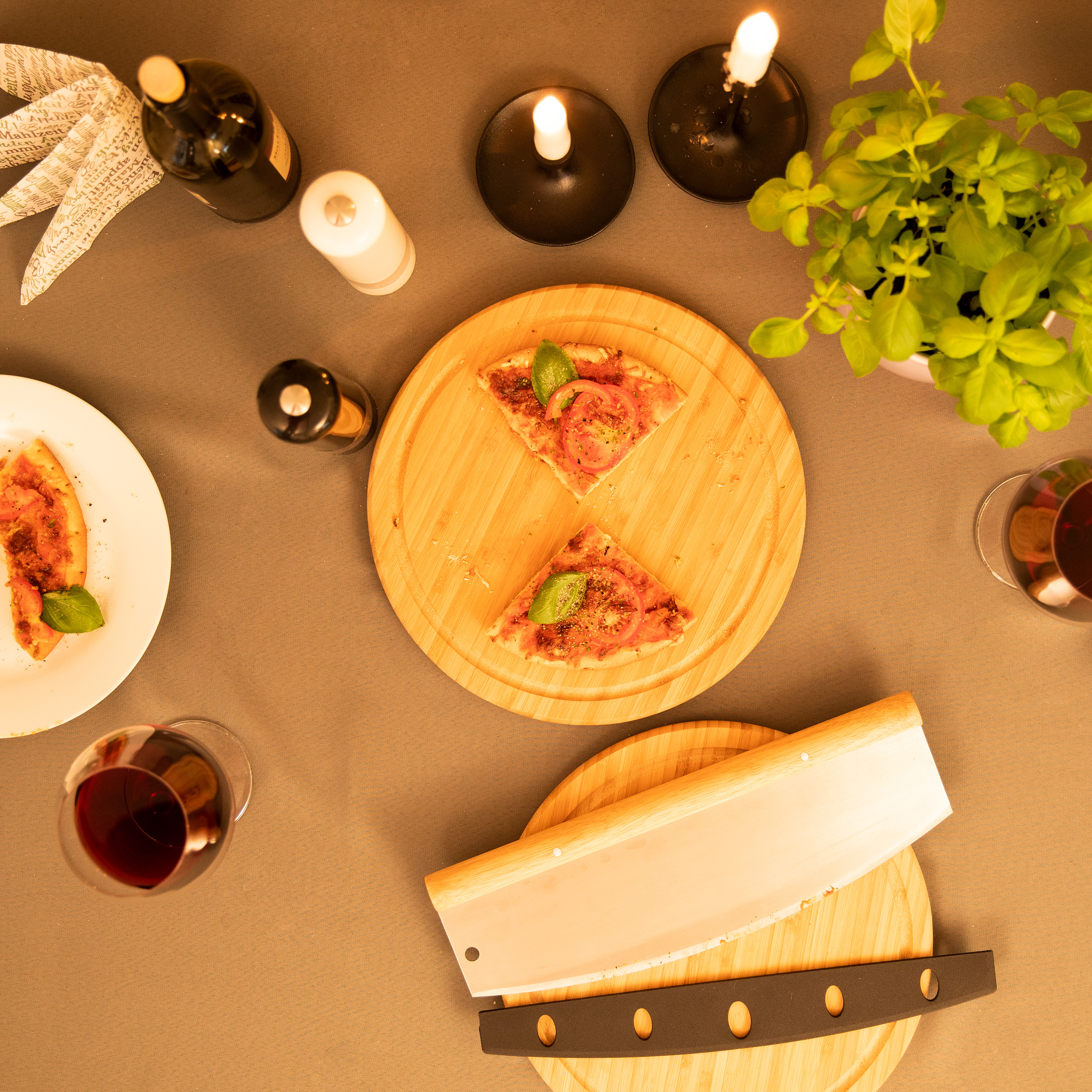 Draufsicht auf einen Esstisch mit Pizzastücken, Gläsern Wein, Kerzen, einer Weinflasche und einer großen Klinge.