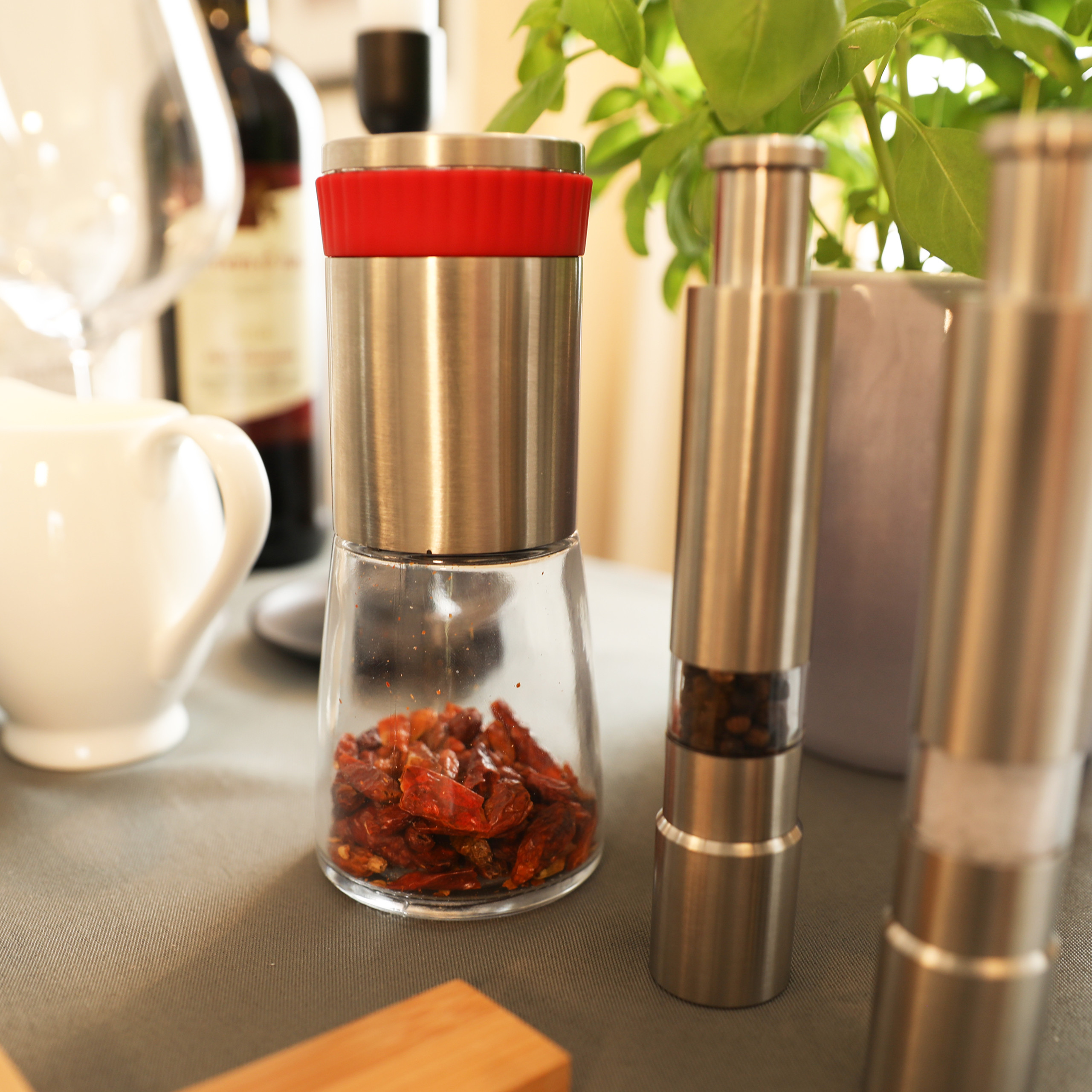 Eine Chili-Mühle mit roter Kappe und zwei Metallmühlen stehen auf einer Küchentheke, im Hintergrund Weinflasche und Basilikum.