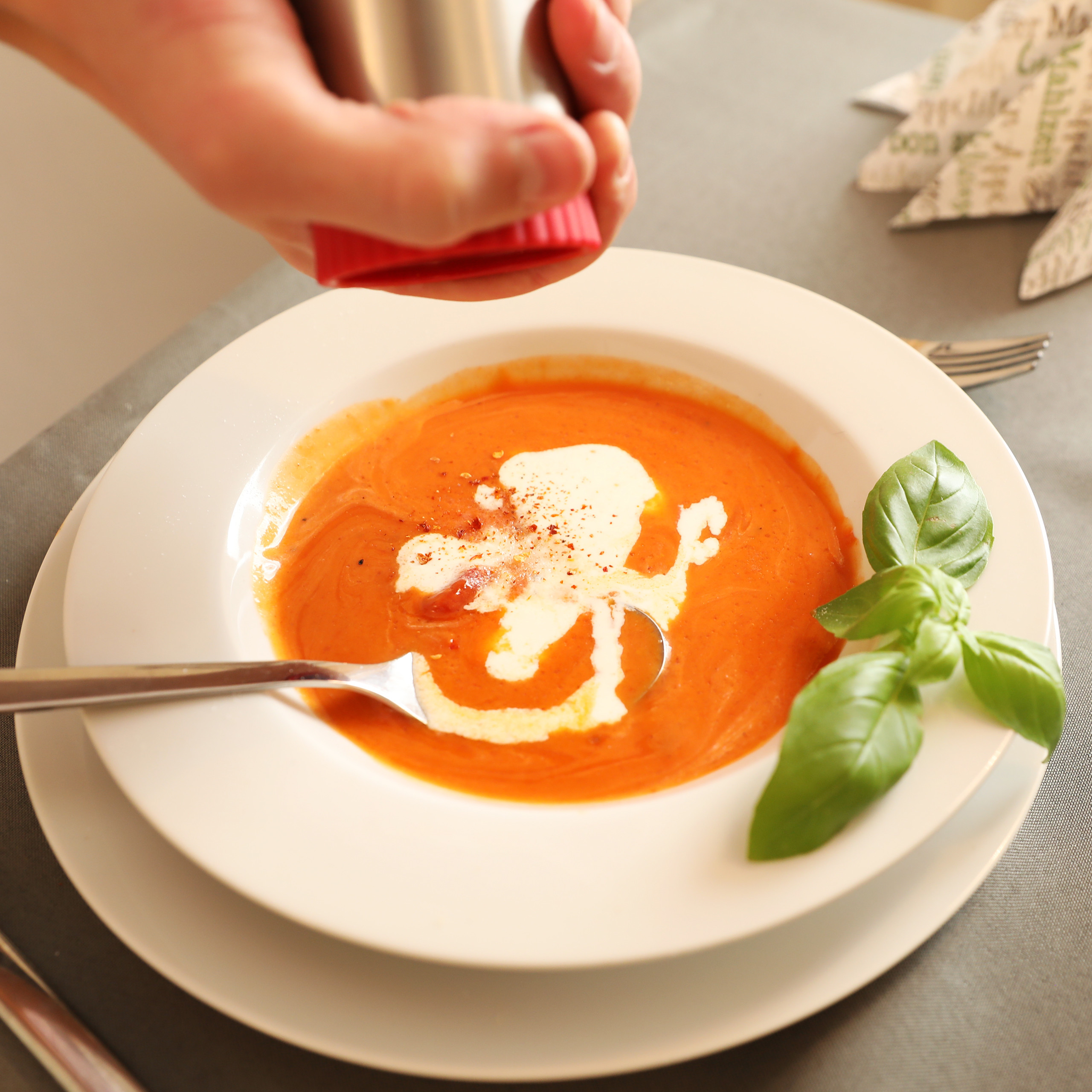 Eine Hand würzt eine cremige Tomatensuppe mit Sahne und Basilikum in einem weißen Teller.