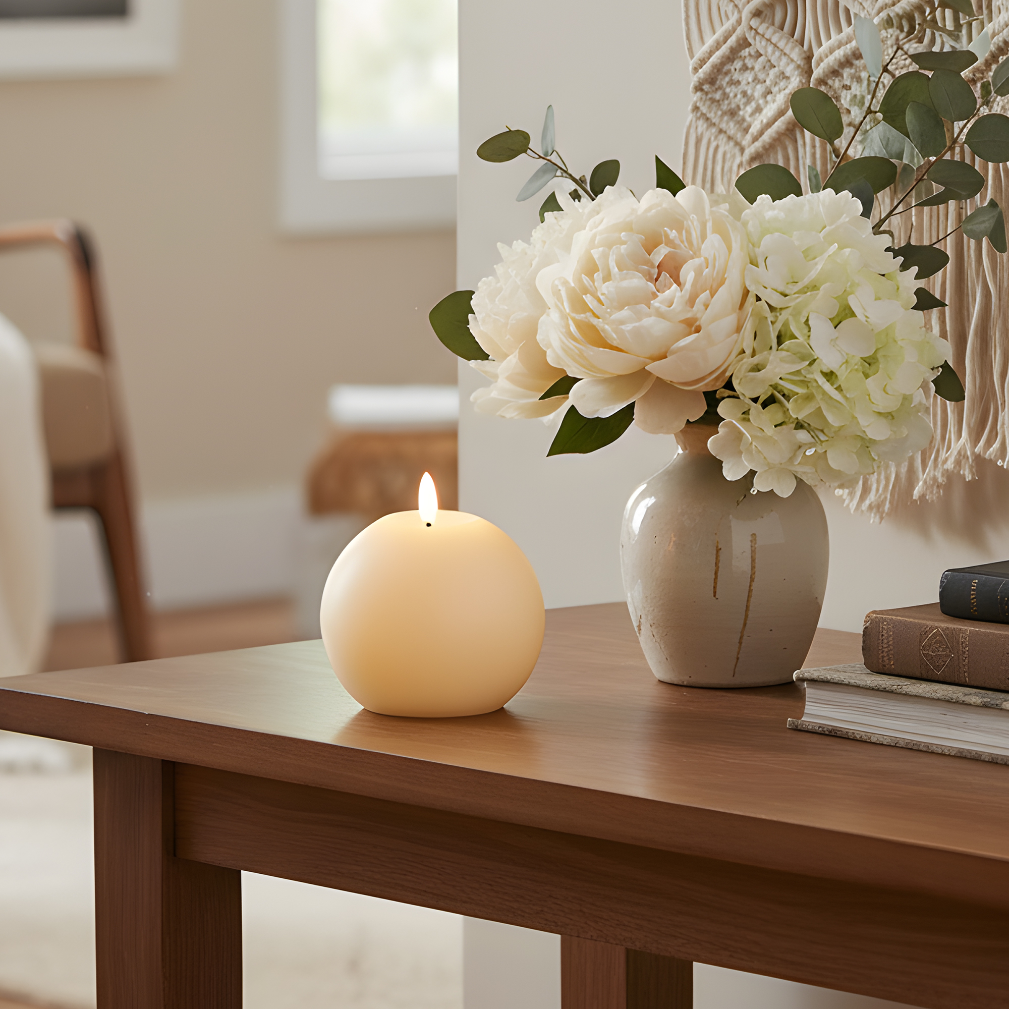 A lit spherical candle, flowers in a vase, and books on a wooden cabinet.