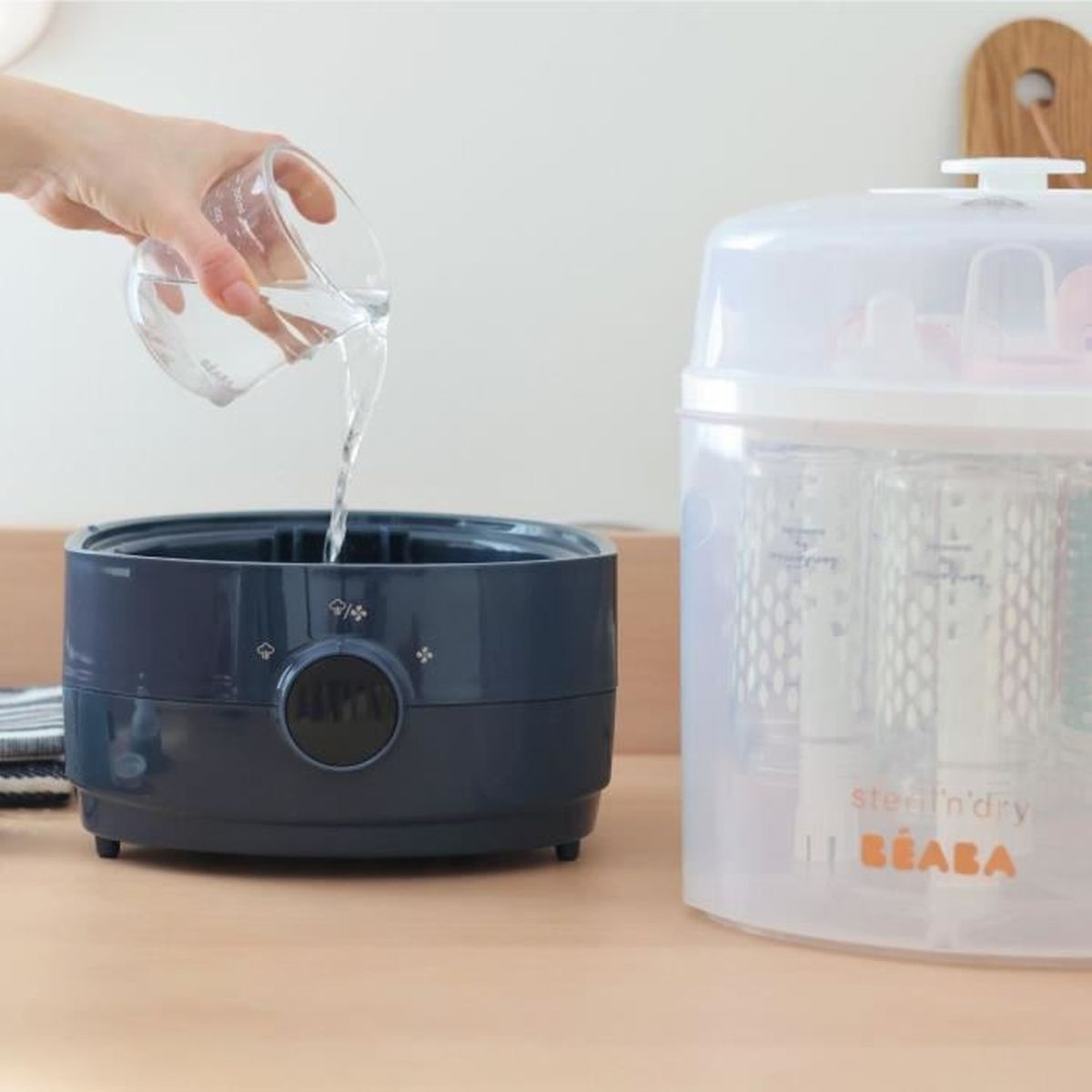 Jemand gießt Wasser aus einem Messbecher in einen blauen Behälter neben einem Sterilisator.