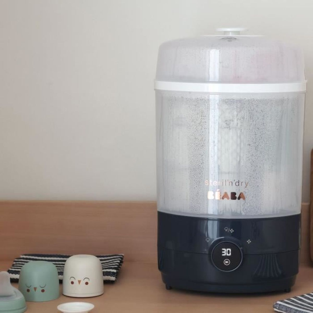 Ein Beaba-Sterilisator mit digitalem Timer steht auf einer Holztheke, daneben zwei kleine Tassen.