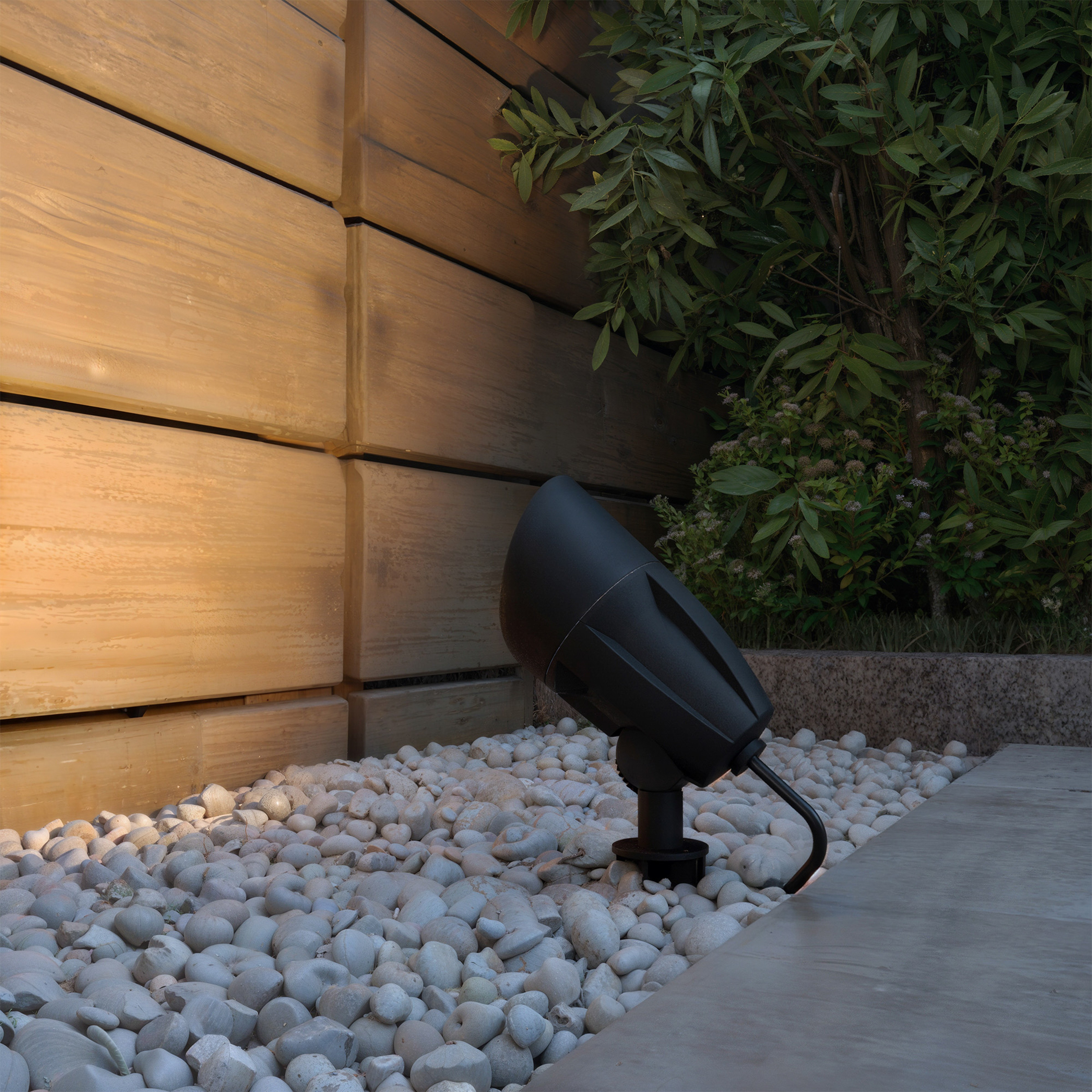 Een zwarte tuinspot staat in witte kiezelstenen voor een houten wand en groene planten.