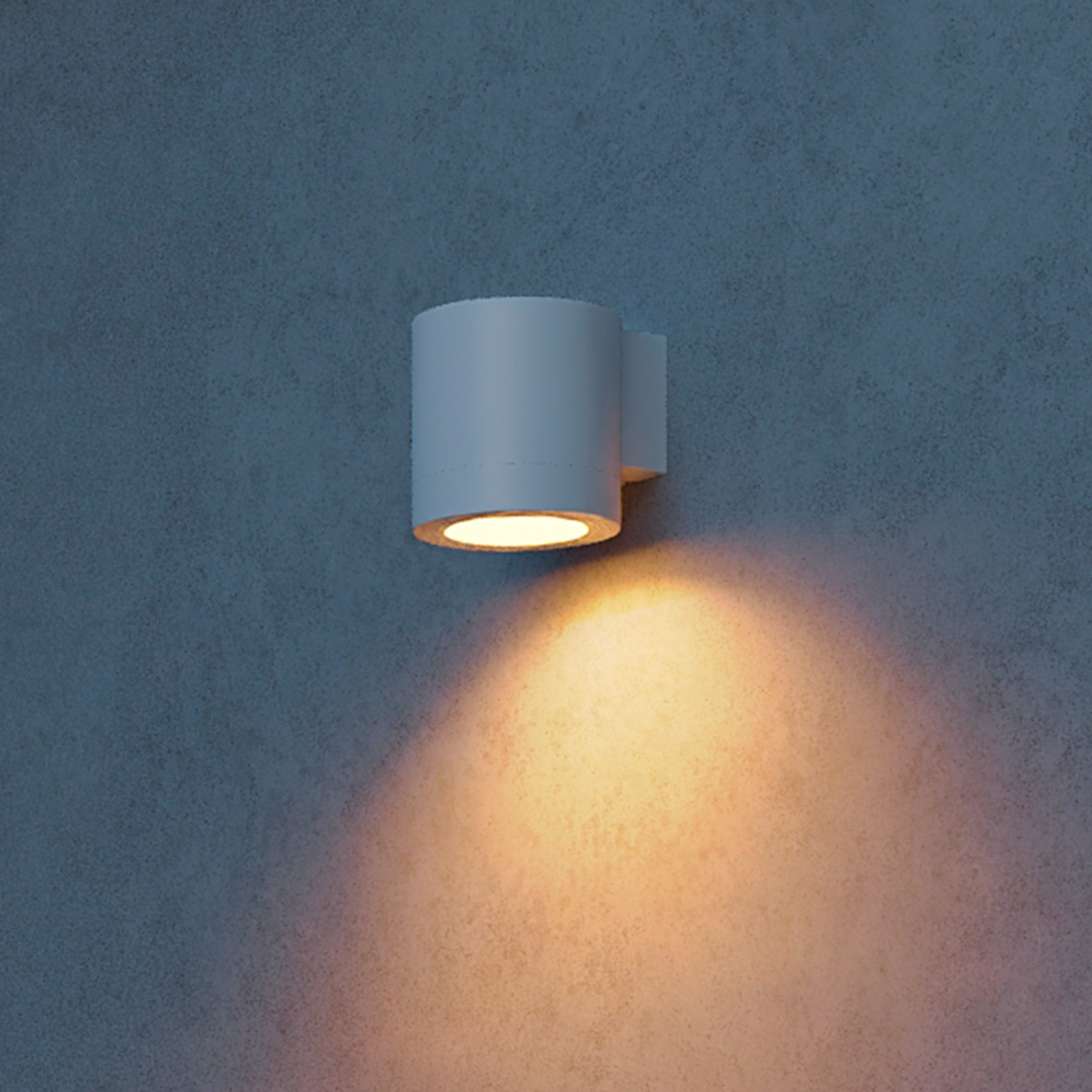 Een cilindrische witte wandlamp op een grijze muur werpt een warm geel licht naar beneden.