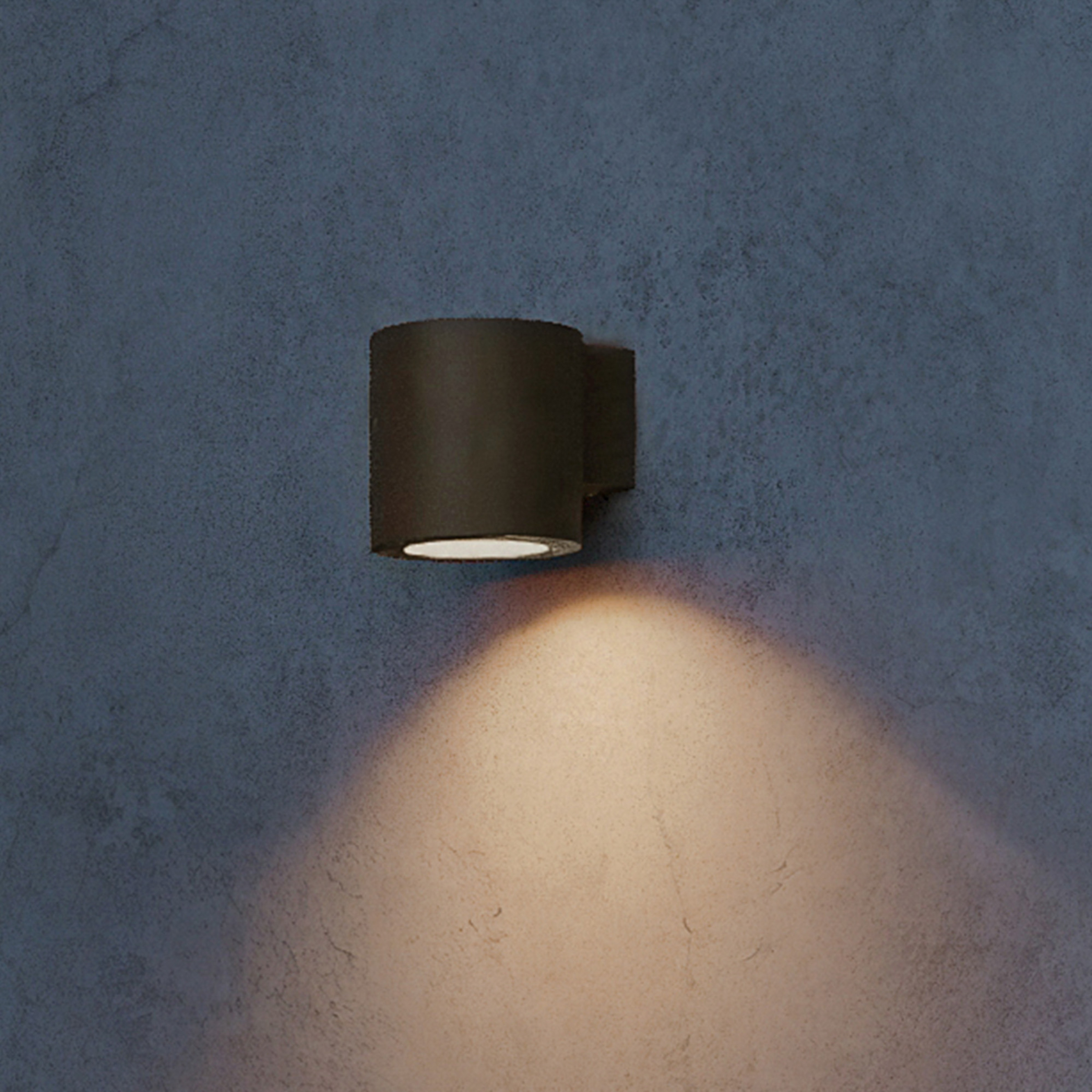 Een zwarte cilindrische wandlamp schijnt warm licht op een grijze muur.