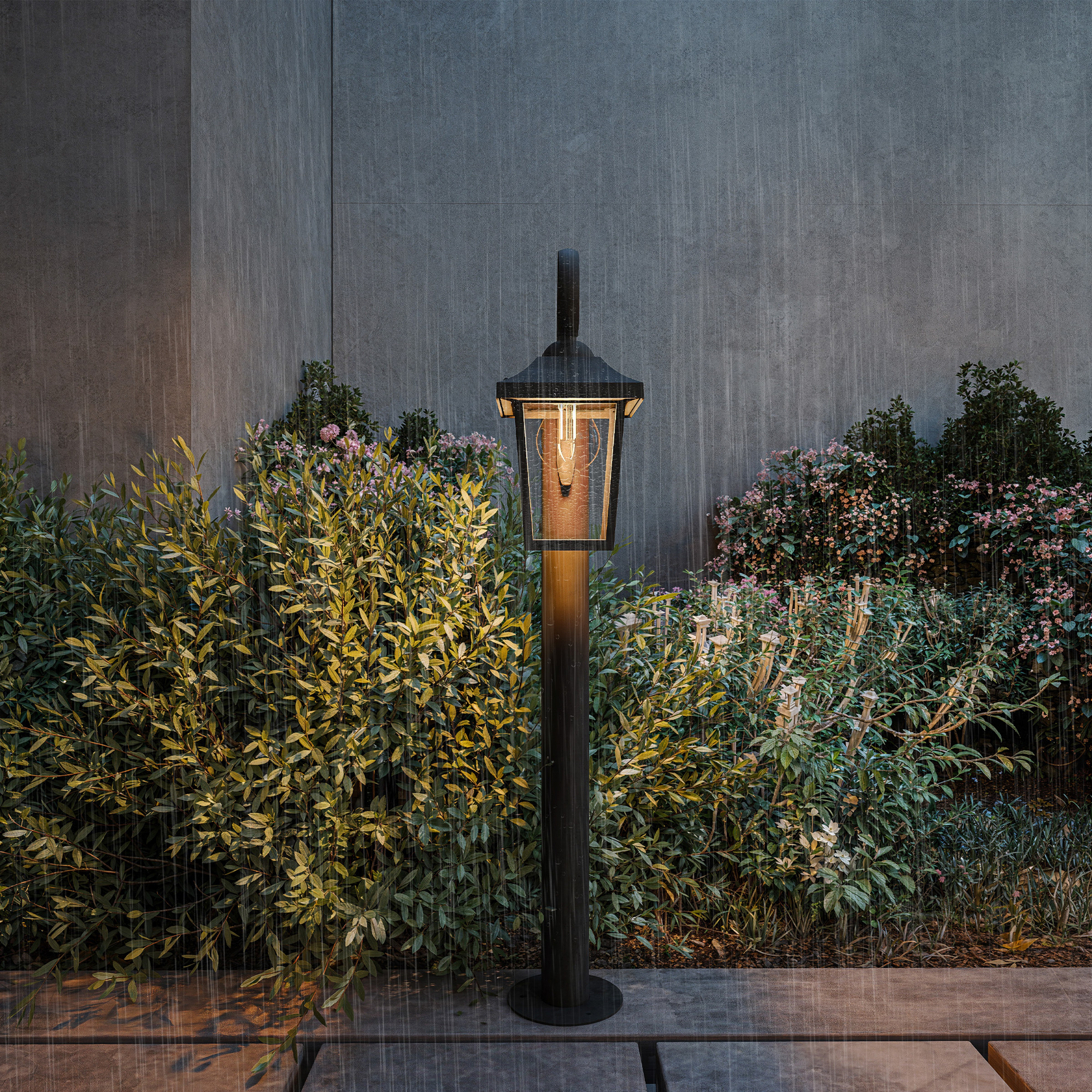 Un lampadaire de jardin noir sous la pluie devant des buissons verts et un mur gris.