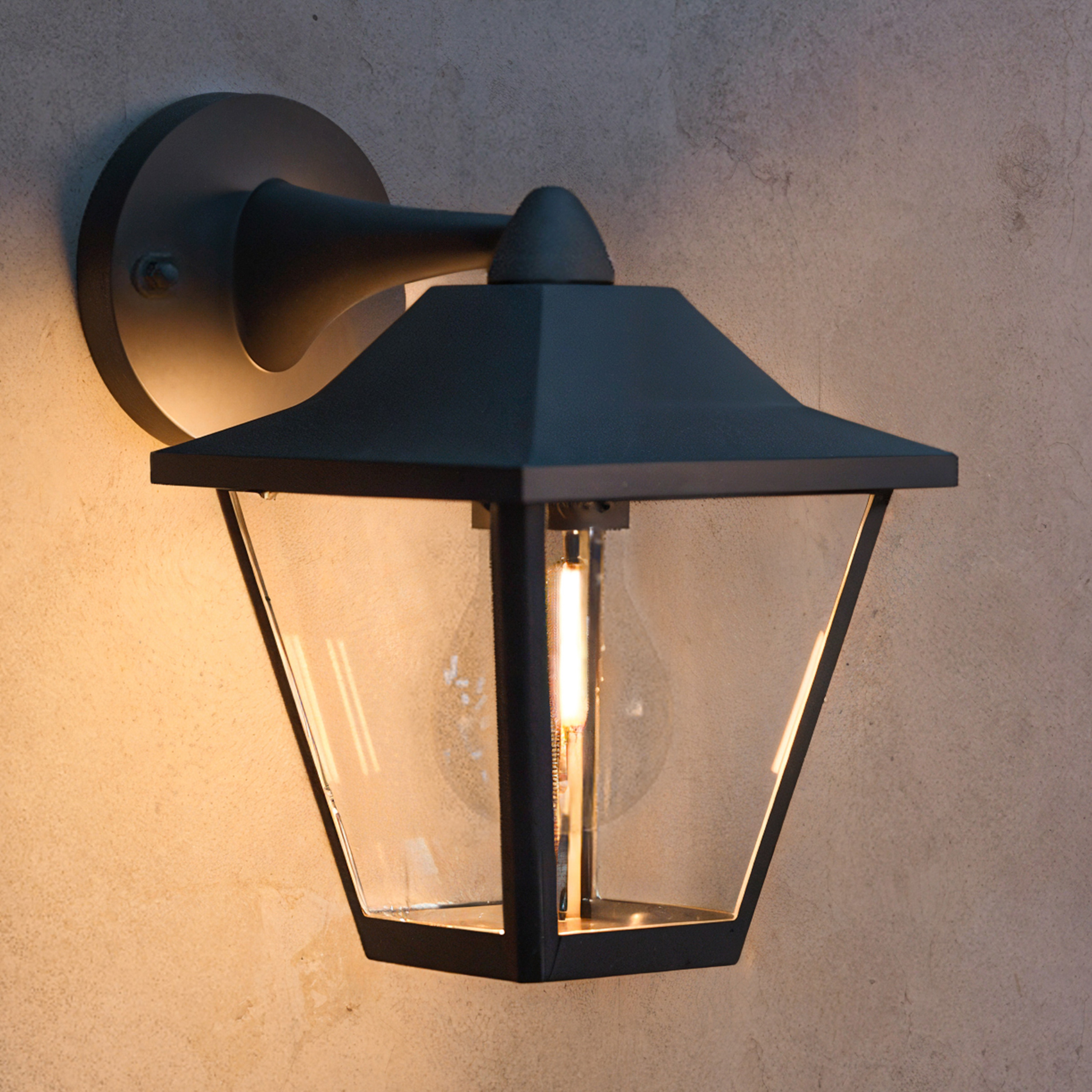 Een zwarte wandlamp met helder glas en warm licht hangt aan een getextureerde muur.