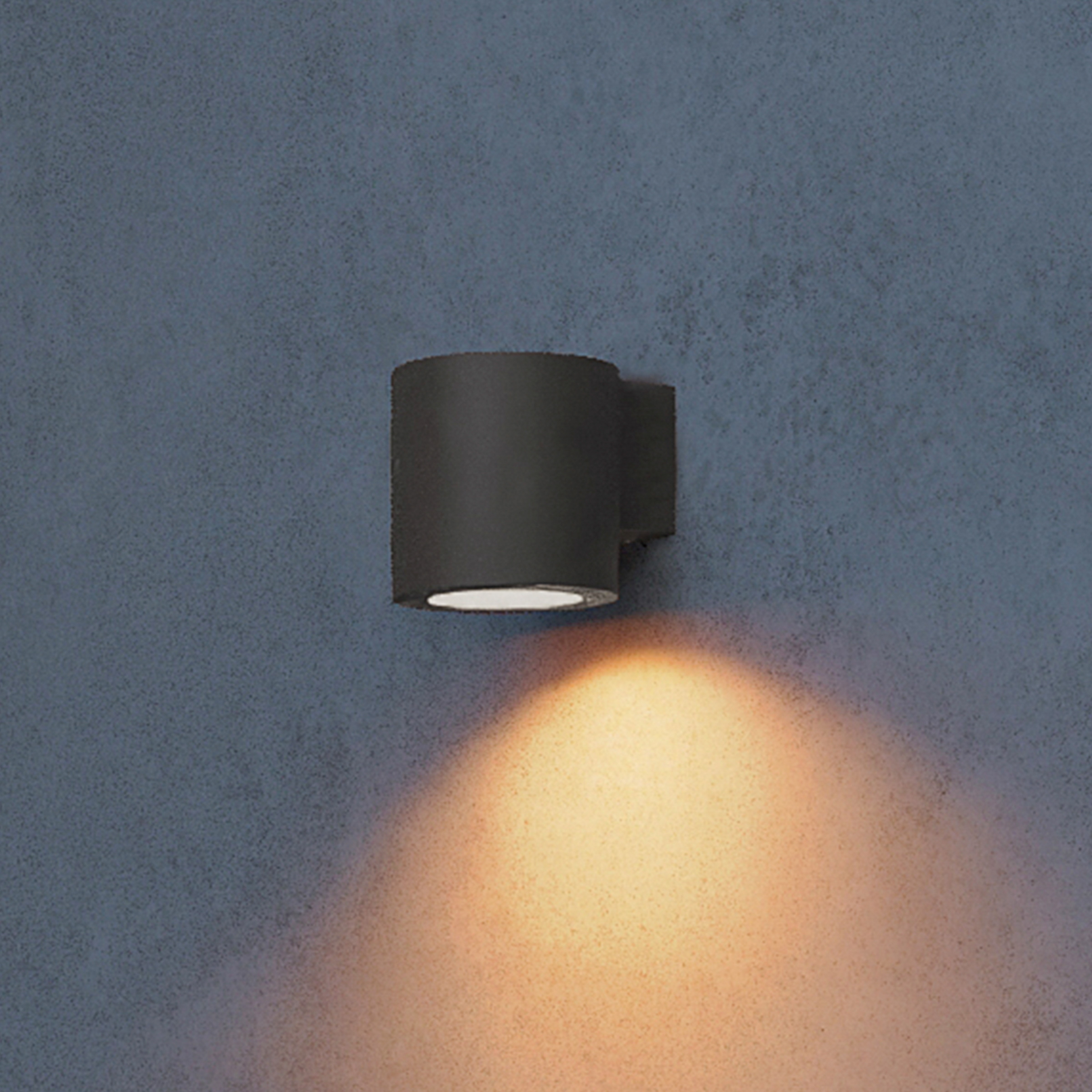 Een zwarte cilindrische wandlamp schijnt warm licht naar beneden op een grijze muur.