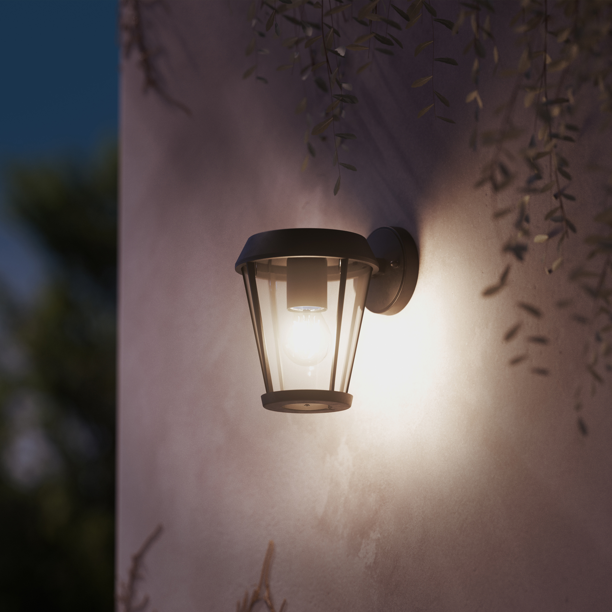 Een verlichte wandlamp aan een roze buitenmuur in de schemering, met hangende planten op de voorgrond.