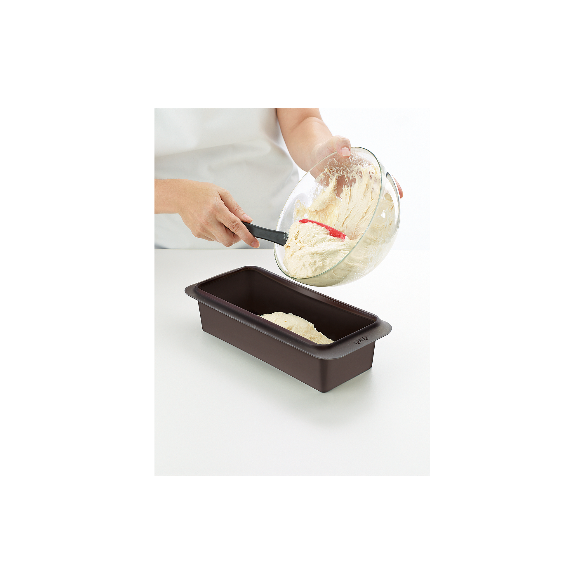 A person pours dough from a glass bowl into a brown loaf pan on a white table using a spatula.