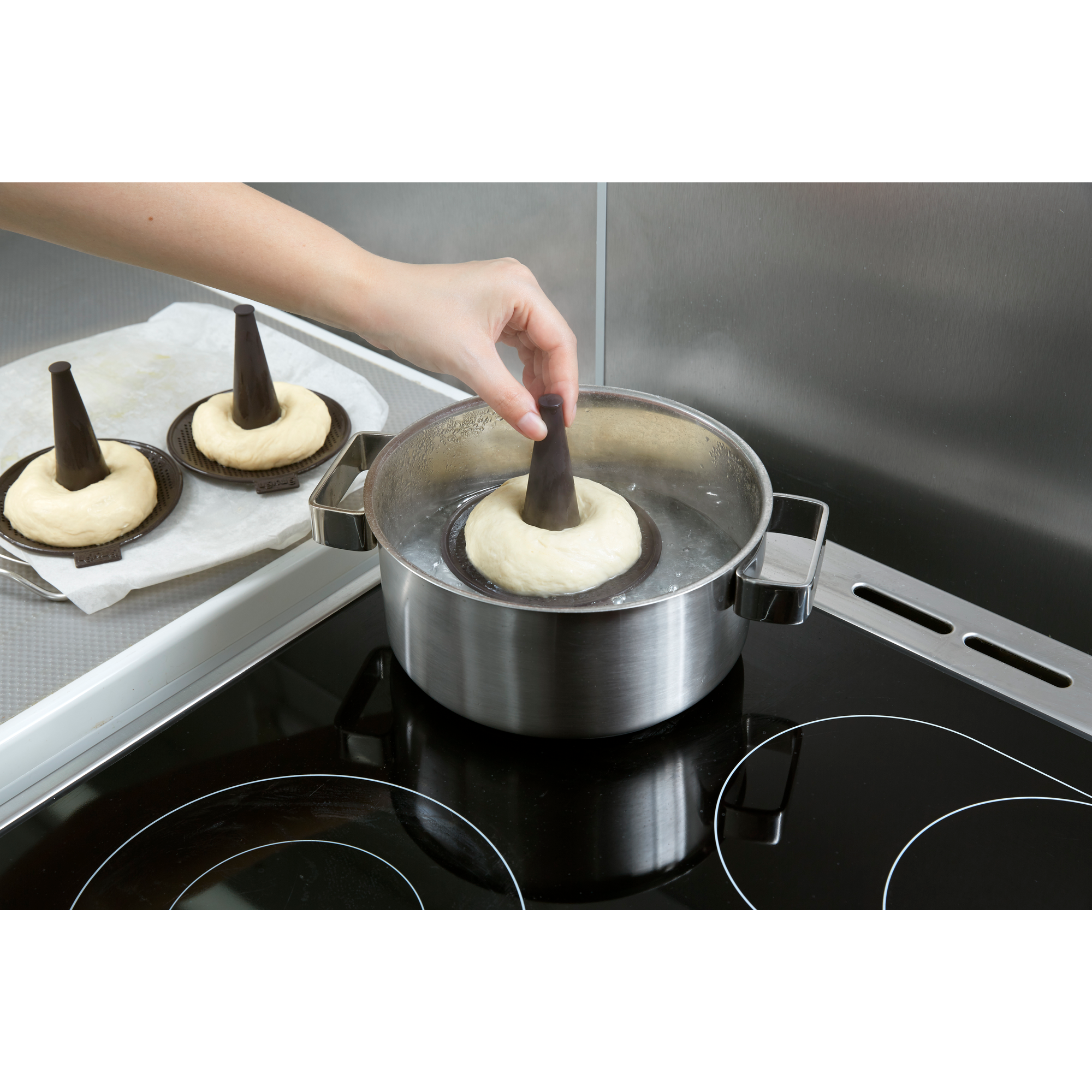 A hand lowers a raw bagel on a mold holder into a pot of boiling water on a stove.