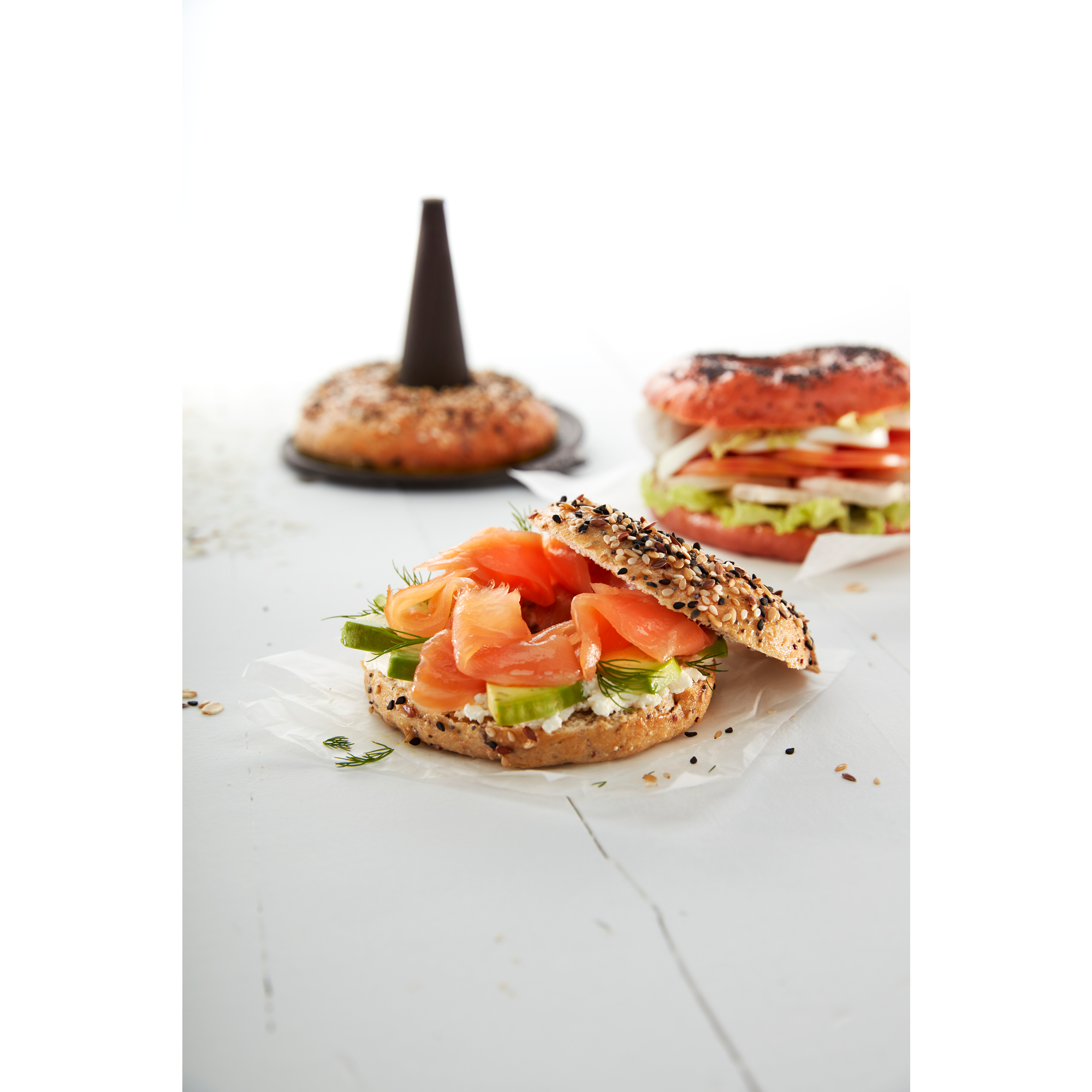 A seeded bagel topped with smoked salmon, cream cheese, avocado, and dill on a white table.