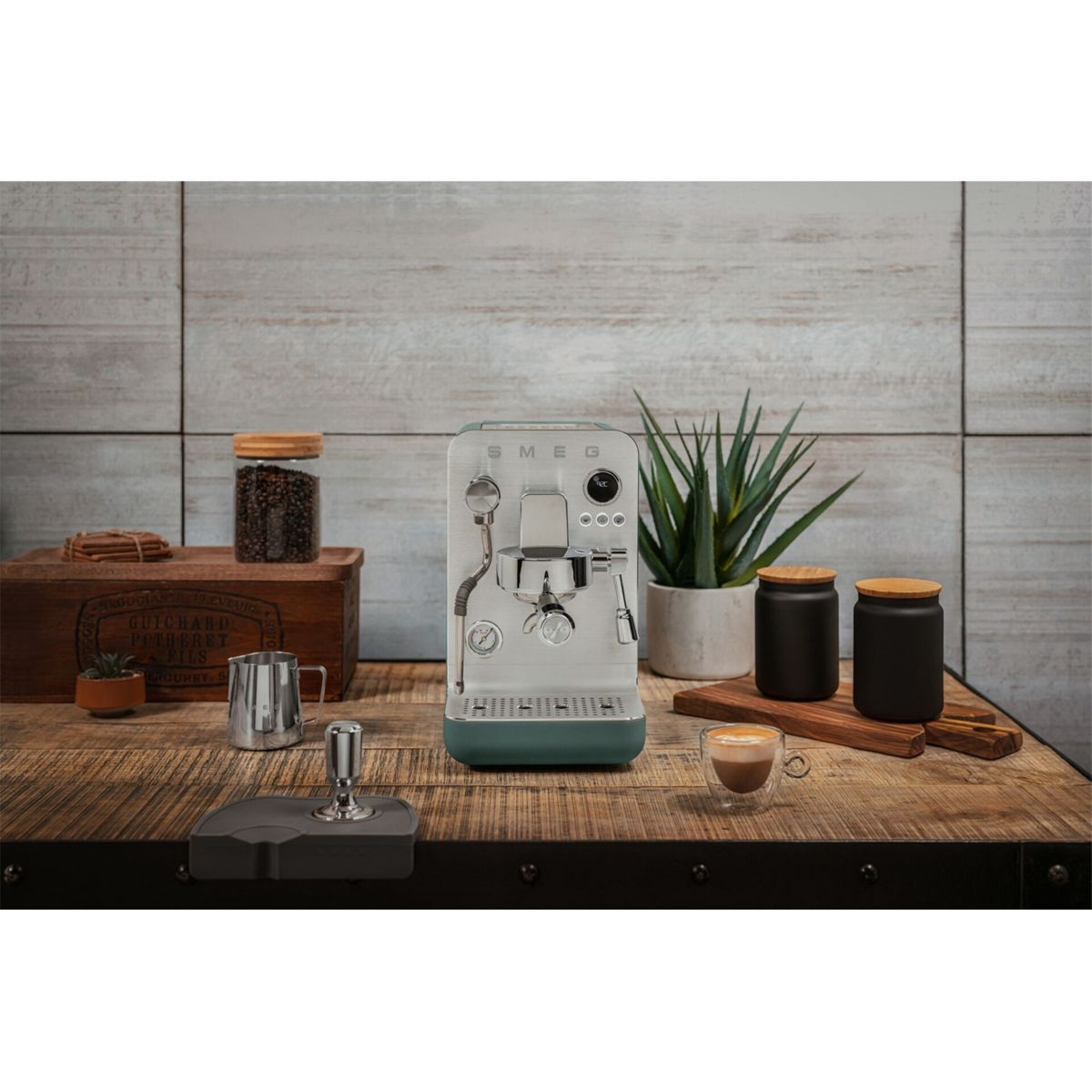 Una máquina de espresso verde sobre una mesa de madera, rodeada de botes de café, una planta y una taza de café.