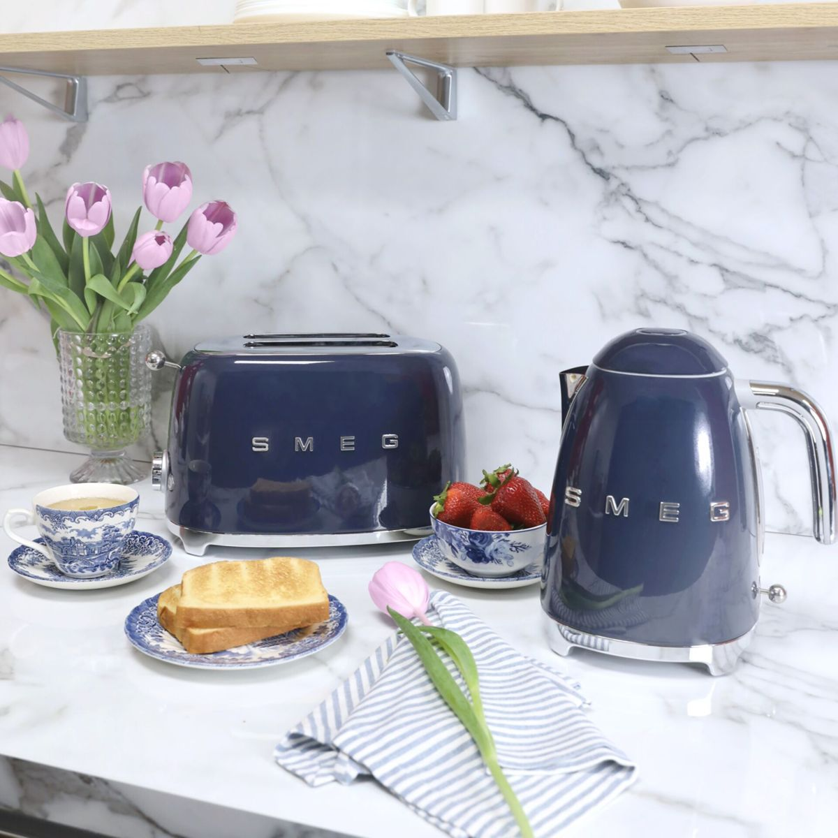Tostapane e bollitore Smeg blu su un piano di marmo con toast, fragole e tulipani rosa.