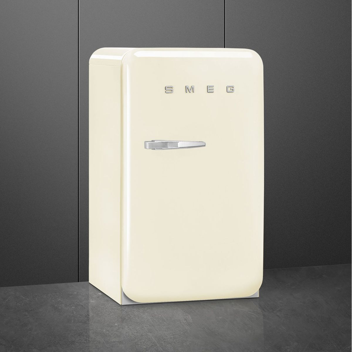 Una mininevera retro crema SMEG con tirador plateado sobre superficie oscura frente a una pared gris.