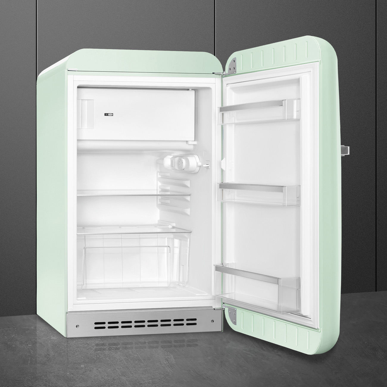 Un refrigerador retro verde menta abierto muestra estantes y compartimentos blancos vacíos contra una pared oscura.