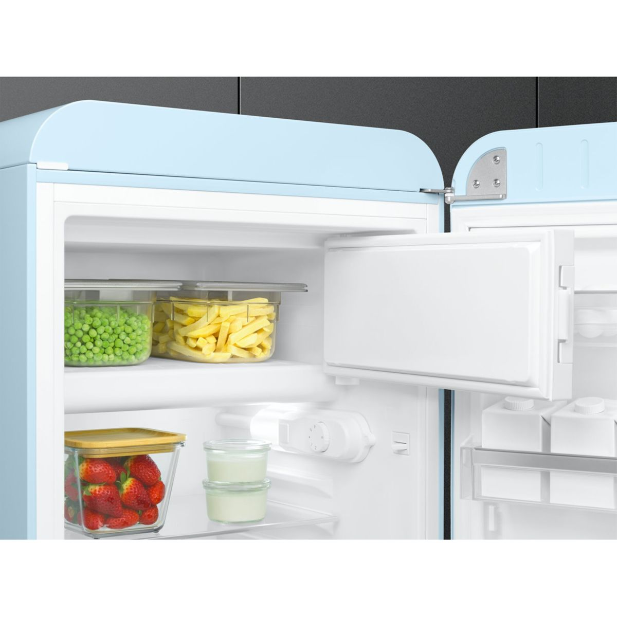 Un refrigerador abierto de color azul claro lleno de recipientes de vidrio con guisantes, patatas fritas, fresas y yogur.