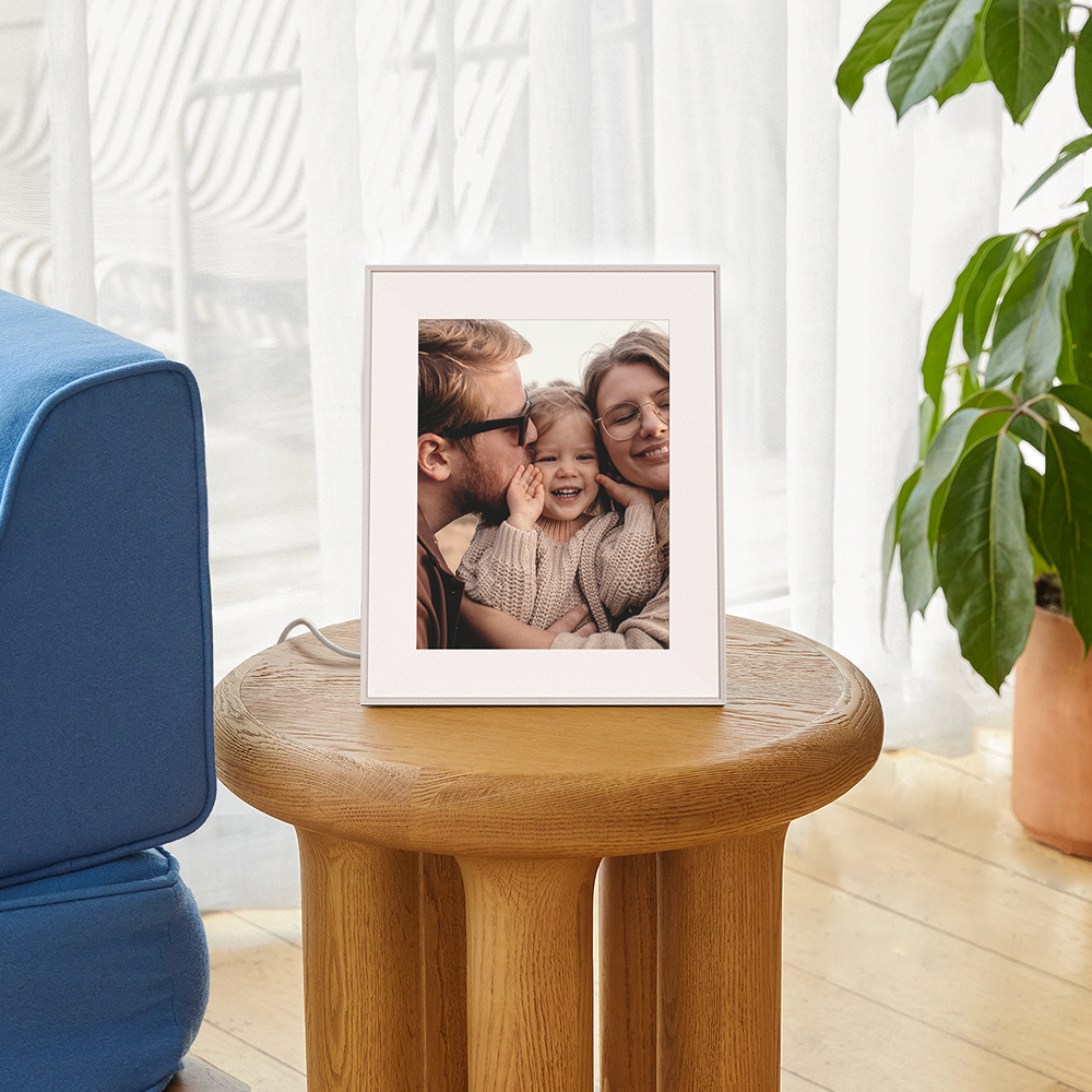 Ein weißer digitaler Bilderrahmen mit einer Familie auf einem runden Holztisch, neben einer blauen Couch und Pflanzen.
