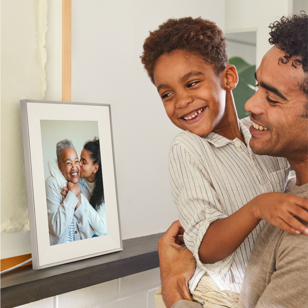 Ein Vater lächelt glücklich, während er seinen Sohn hält, und betrachtet einen digitalen Bilderrahmen mit einer Familie.