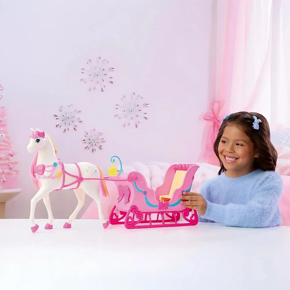 Una niña sonriente con jersey azul juega con un caballo de juguete blanco y un trineo rosa.