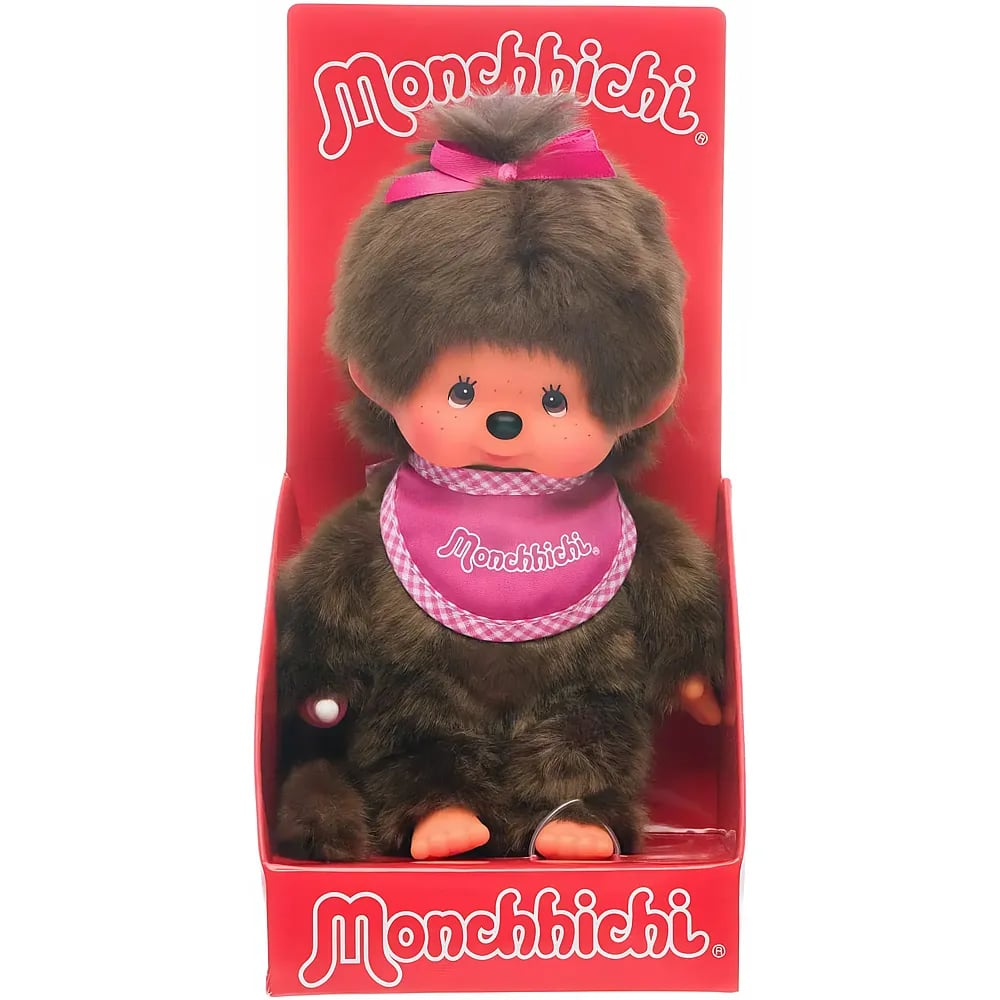 Un muñeco de peluche Monchhichi marrón con babero rosa y lazo, dentro de su caja roja.