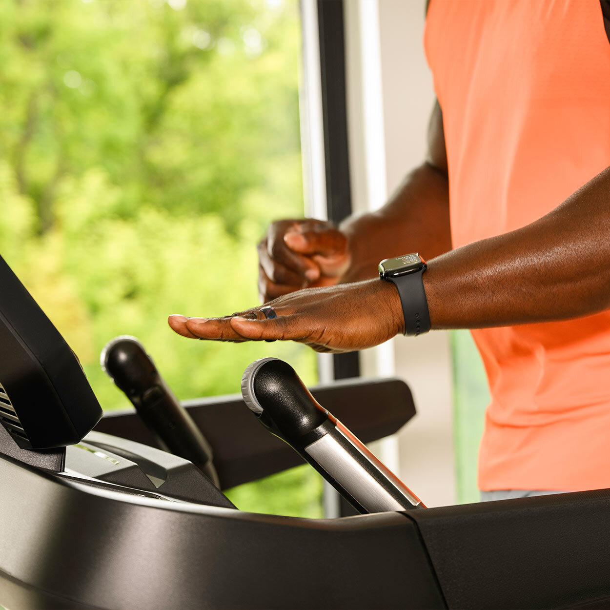Ein Mann im orangen Tanktop läuft auf einem Laufband und trägt eine Smartwatch am Handgelenk.