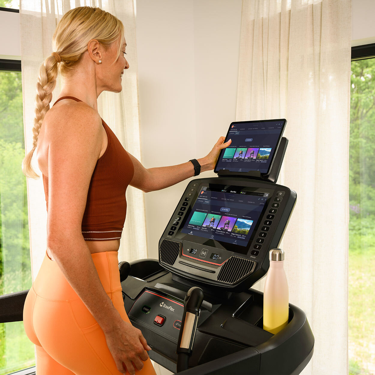Eine blonde Frau in Sportkleidung nutzt ein Laufband mit Bildschirm und Tablet in einem hellen Raum.