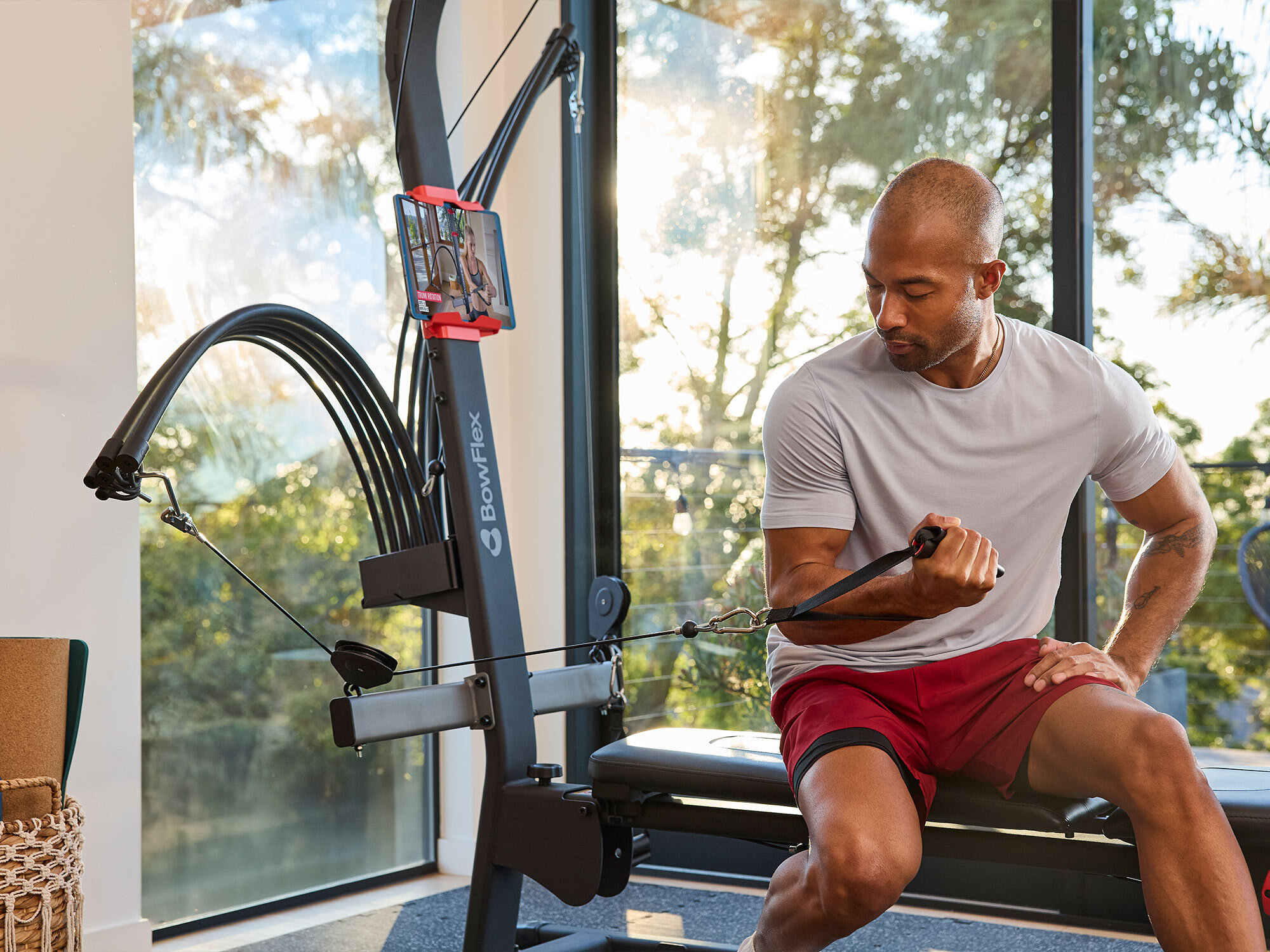 Ein Mann im grauen Shirt und roten Shorts trainiert drinnen an einer schwarzen Bowflex-Maschine mit Tablet.