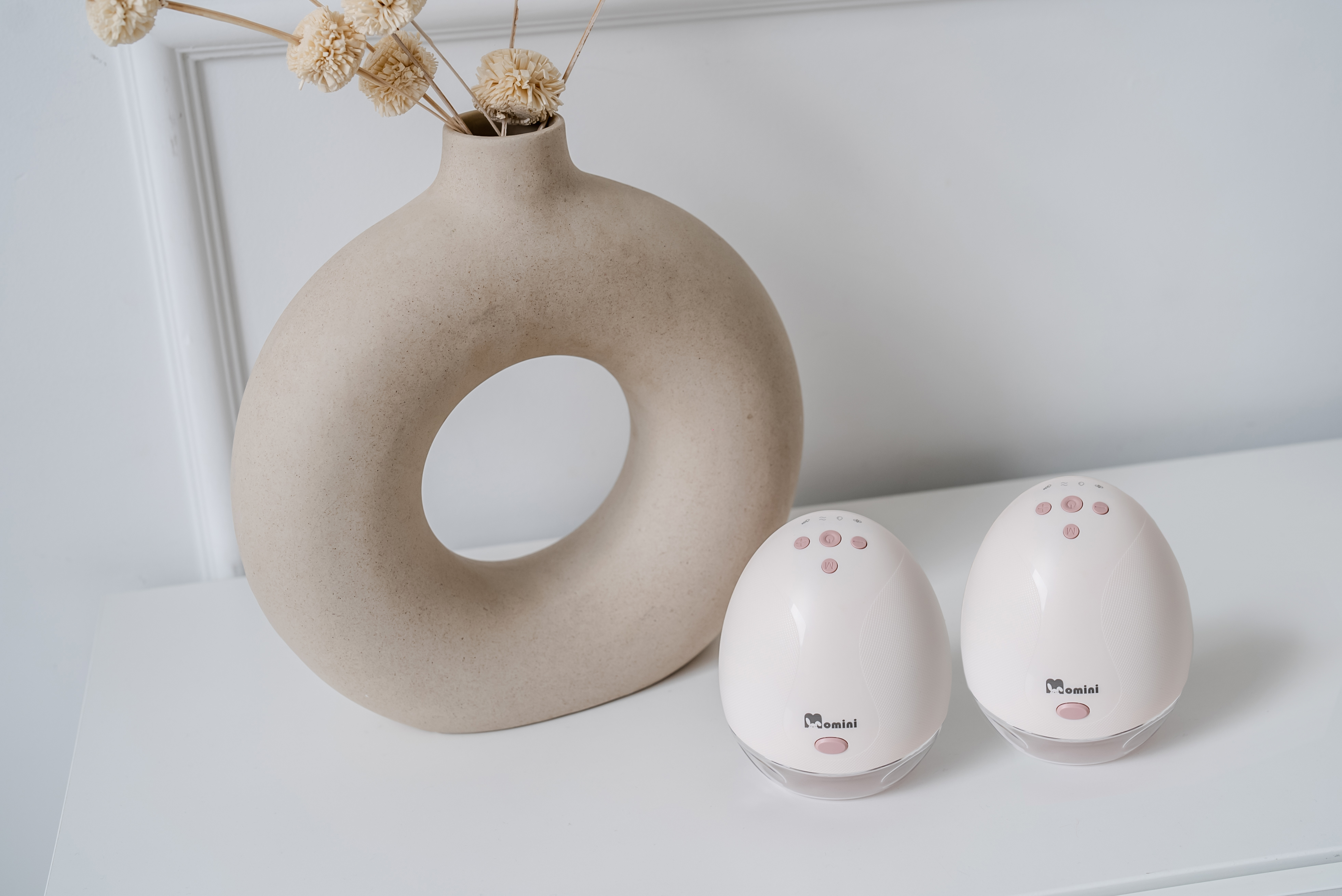 Due tiralatte bianchi Momini su un tavolo bianco accanto a un vaso rotondo beige con fiori secchi.