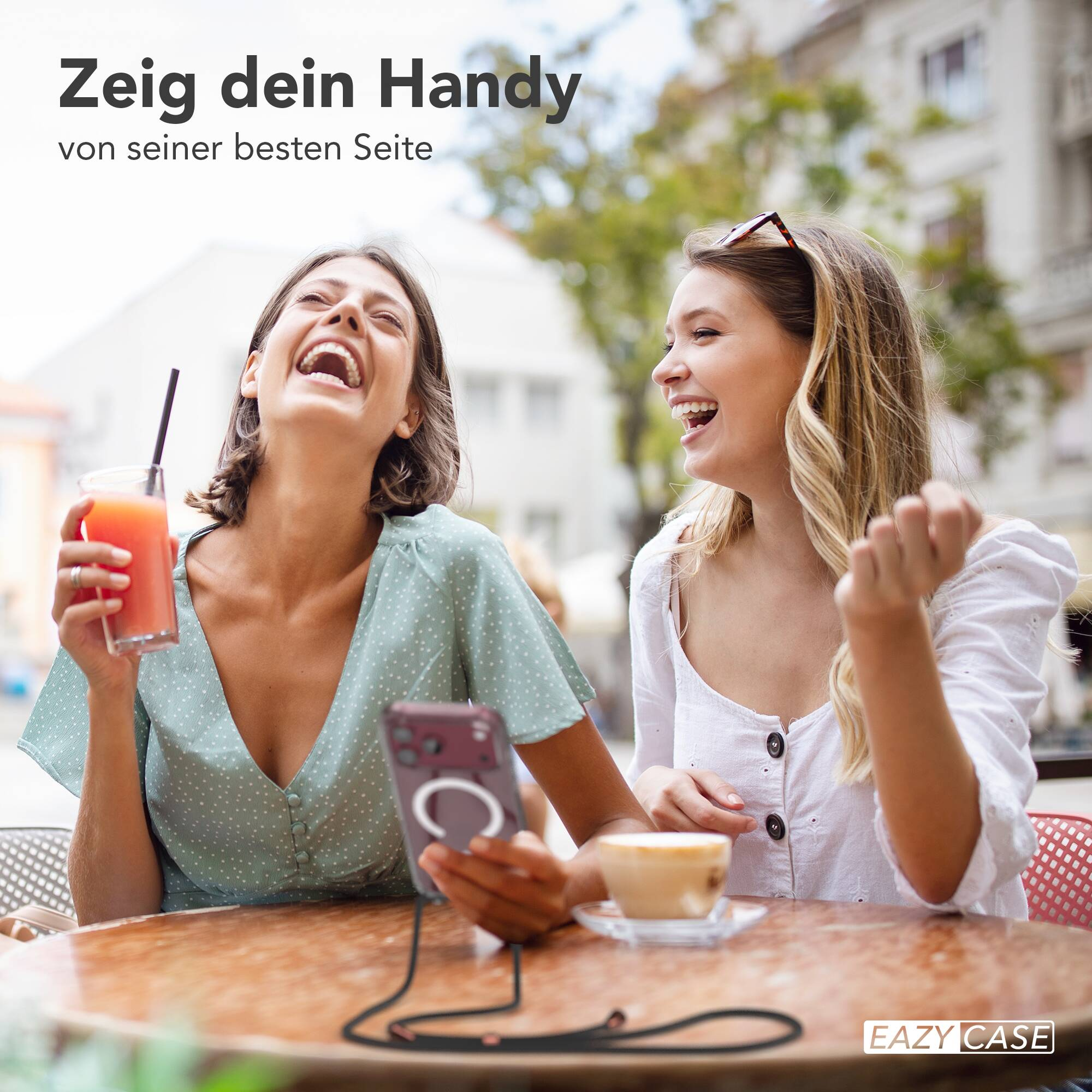 Zwei junge Frauen lachen an einem Tisch in einem Straßencafé mit Getränken und einem Handy.