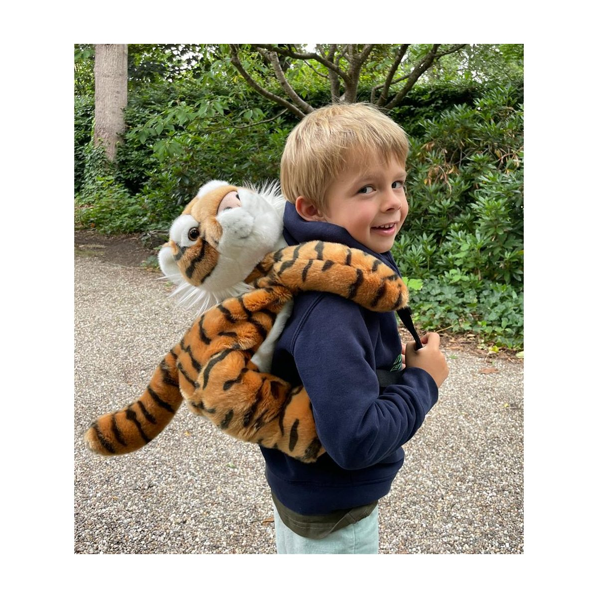 Ein blondes Kind lächelt, während es draußen einen Plüschtiger-Rucksack trägt.