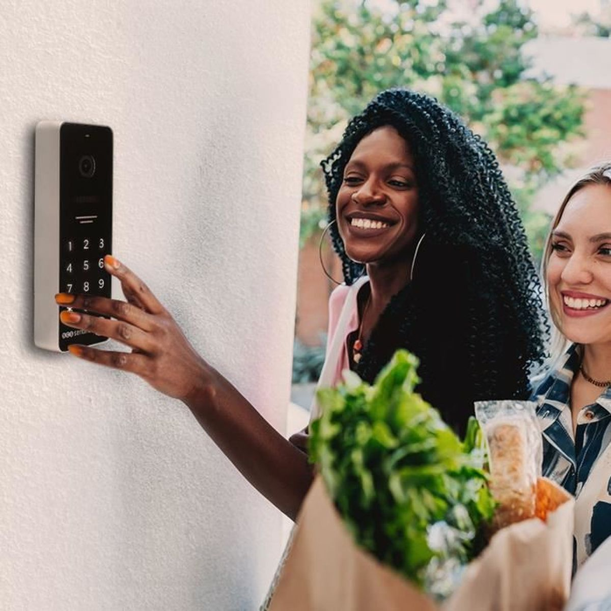 Zwei lächelnde Frauen mit Einkaufstüten tippen draußen einen Code in eine Gegensprechanlage.